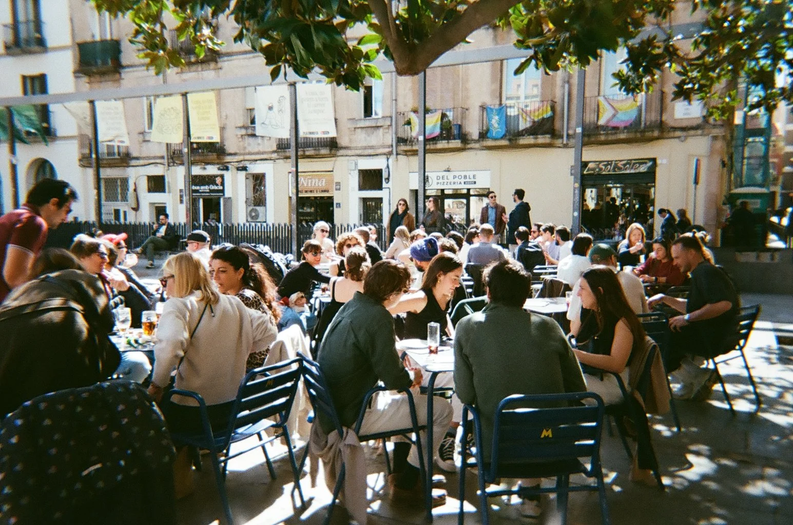 People dining outdoors at a cafe on a sunny day in an urban setting, with buildings in the background.