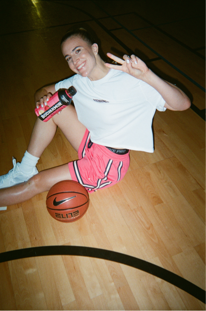 A young woman sitting on a basketball court floor, smiling and holding a pink water bottle, making a peace sign with her right hand, with a Nike basketball nearby.