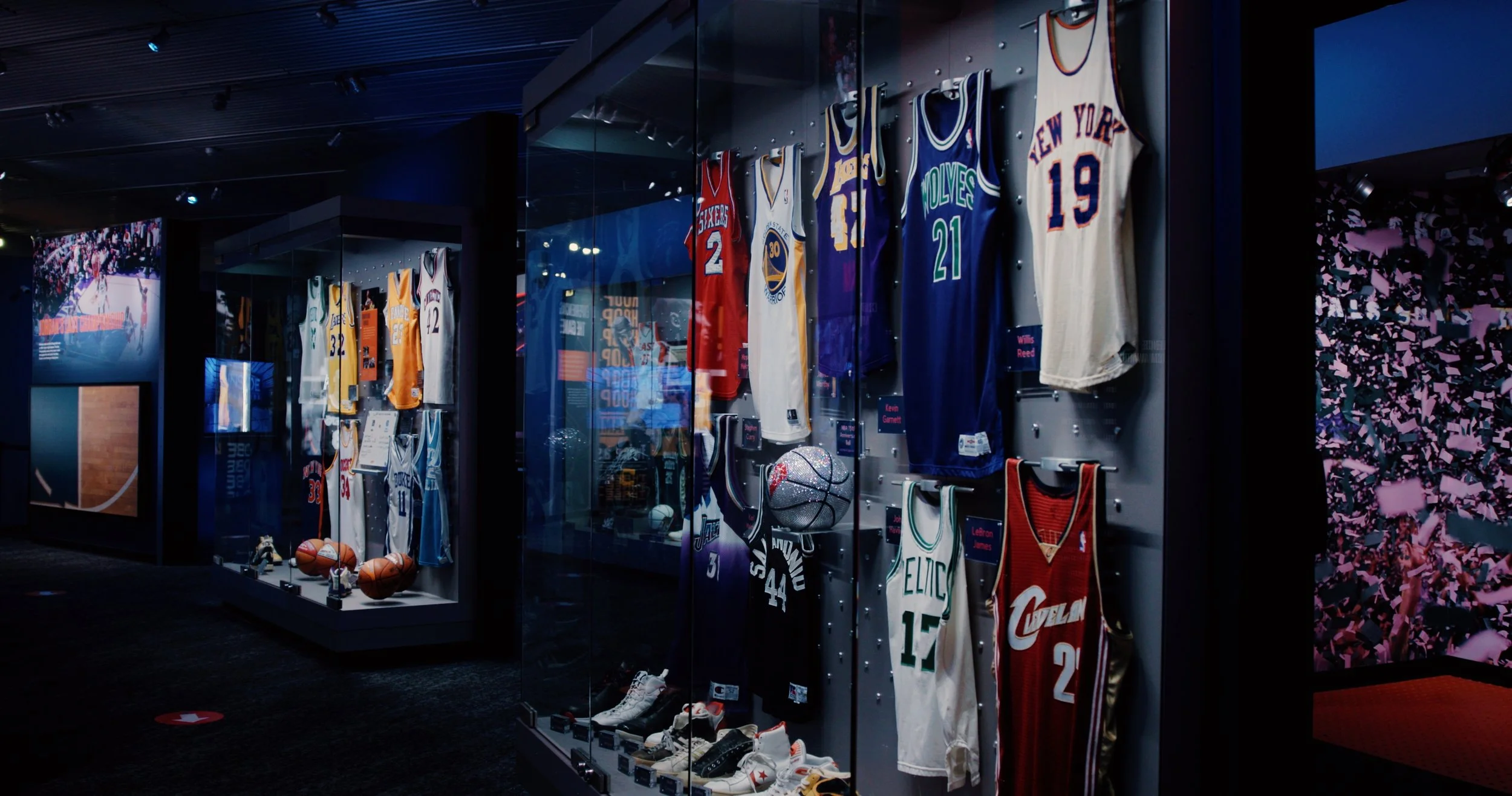Display case with basketball jerseys from various NBA teams, basketballs, and sneakers in a sports museum or exhibit.