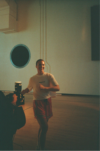 Person standing in a gym or dance studio, smiling, wearing a white t-shirt and red shorts, with a speaker nearby and a circular window in the background.
