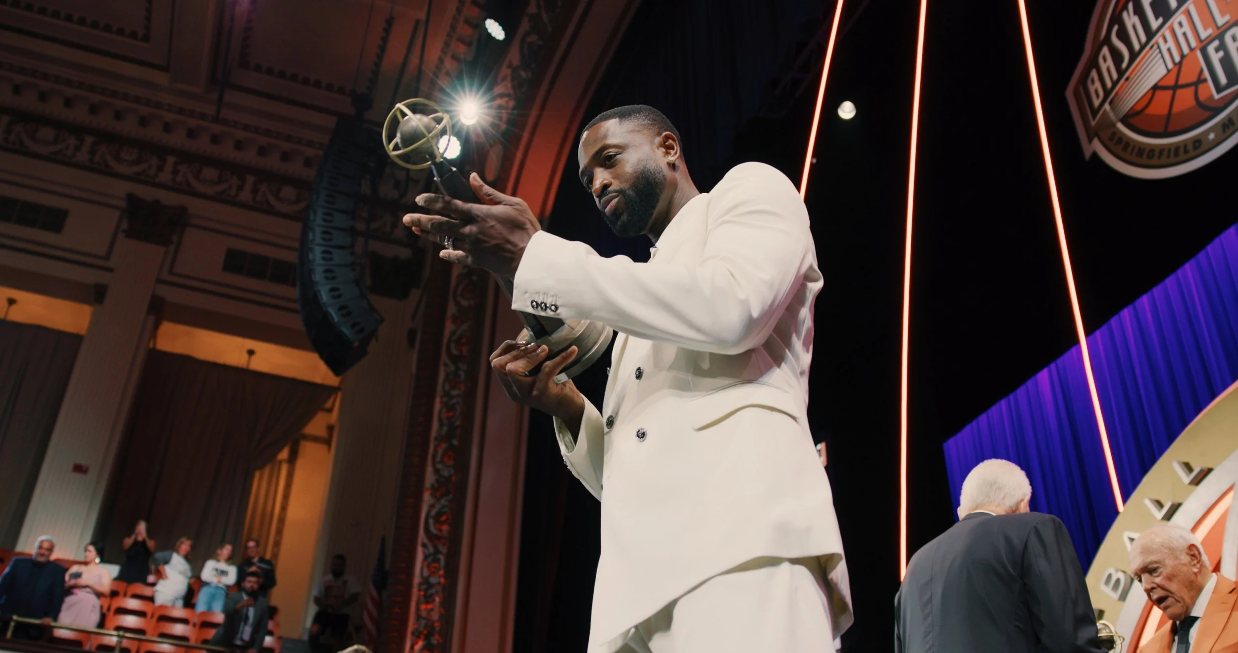 Dwyane wade holding an award on stage at an event with a basketball logo in the background.