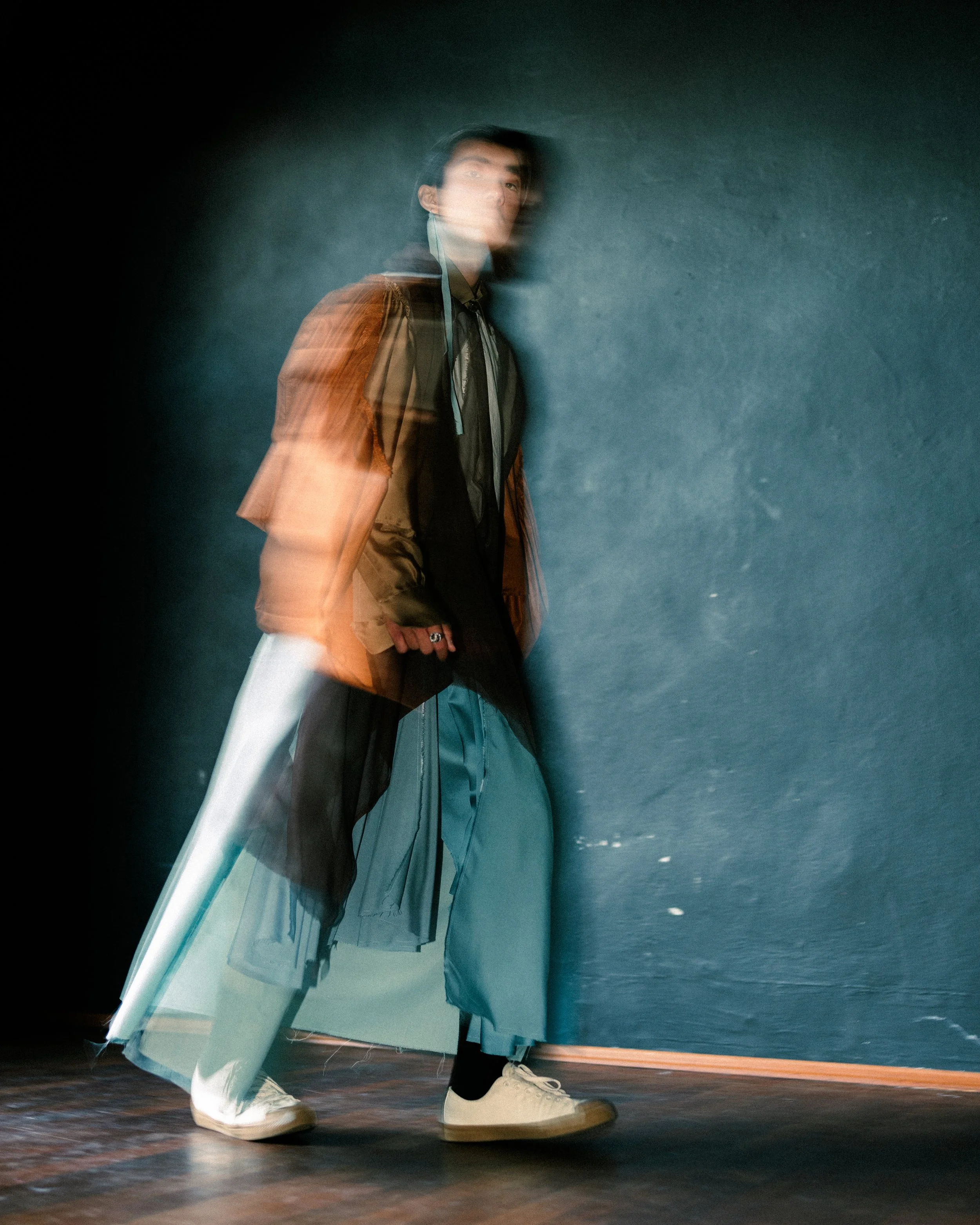 A person standing and leaning against a dark green wall, with motion blur creating ghostly overlapping images of the person in a layered effect.