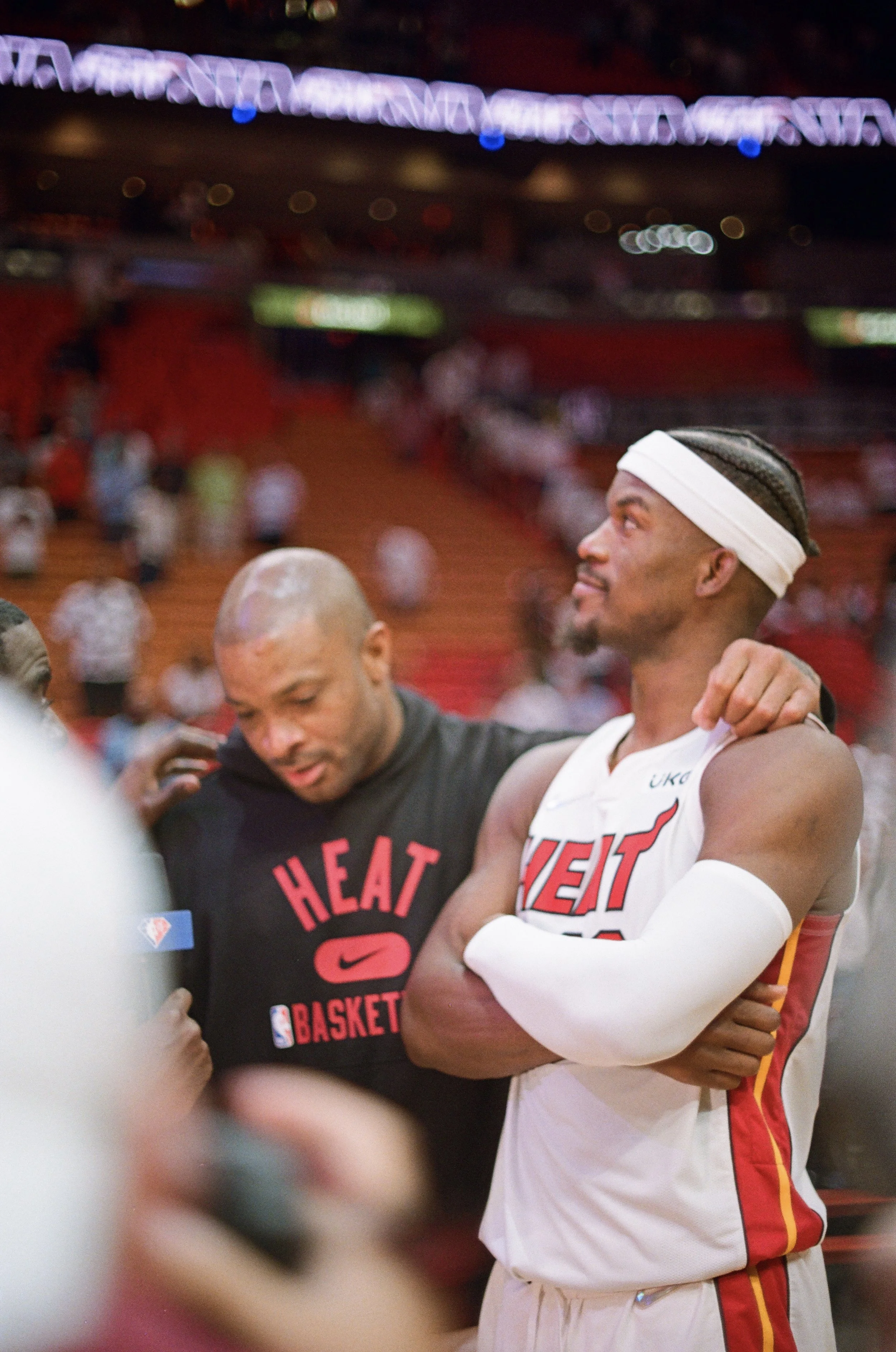Miami Heat basketball player with arms crossed and wearing white wrap, standing next to a teammate in a black shirt, in an arena.