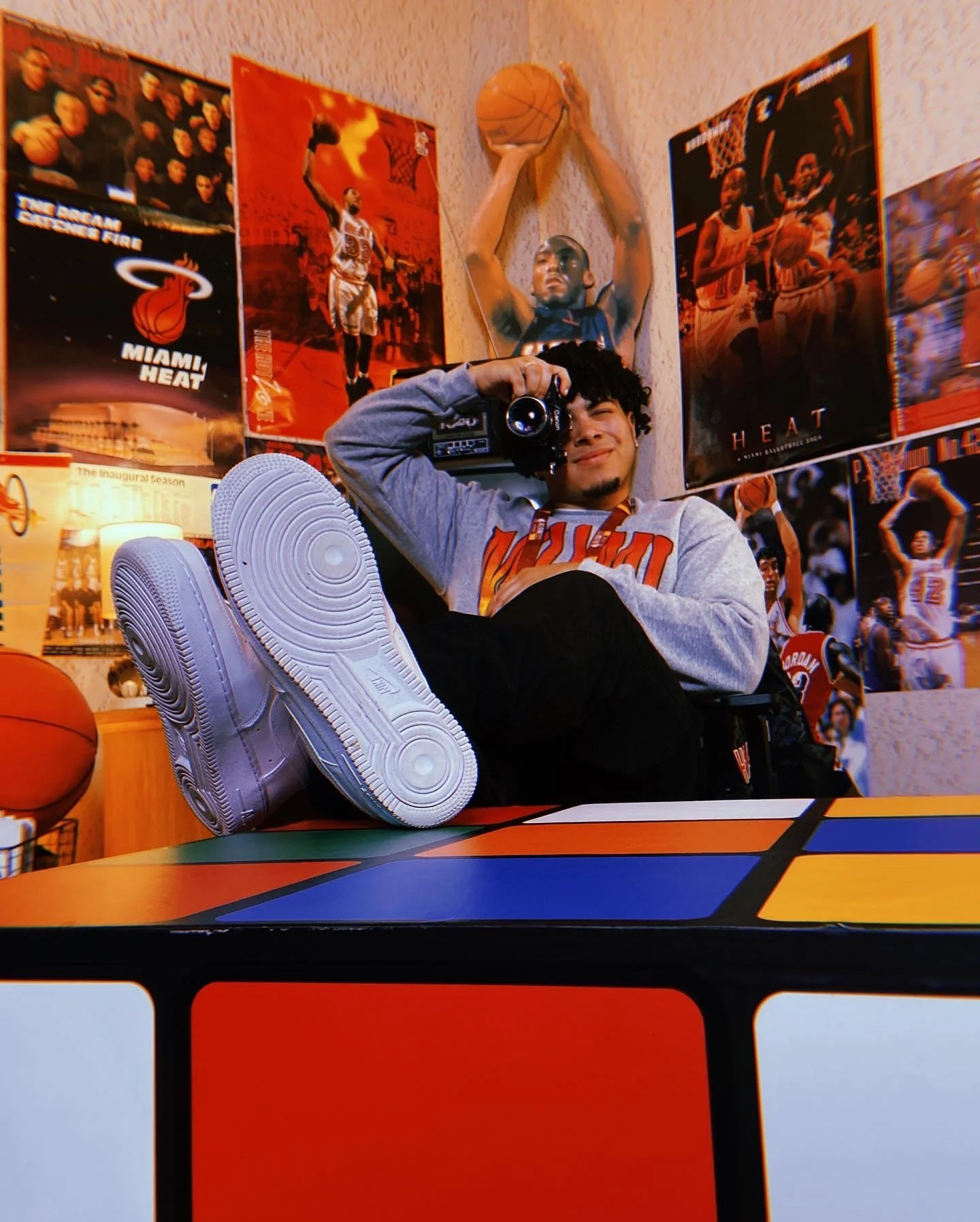 A young man with curly hair is sitting on a chair with his legs stretched out, wearing white sneakers, a gray hoodie, and black pants. He is holding a camera near his face, as if taking a photo. The room is decorated with basketball posters and memor