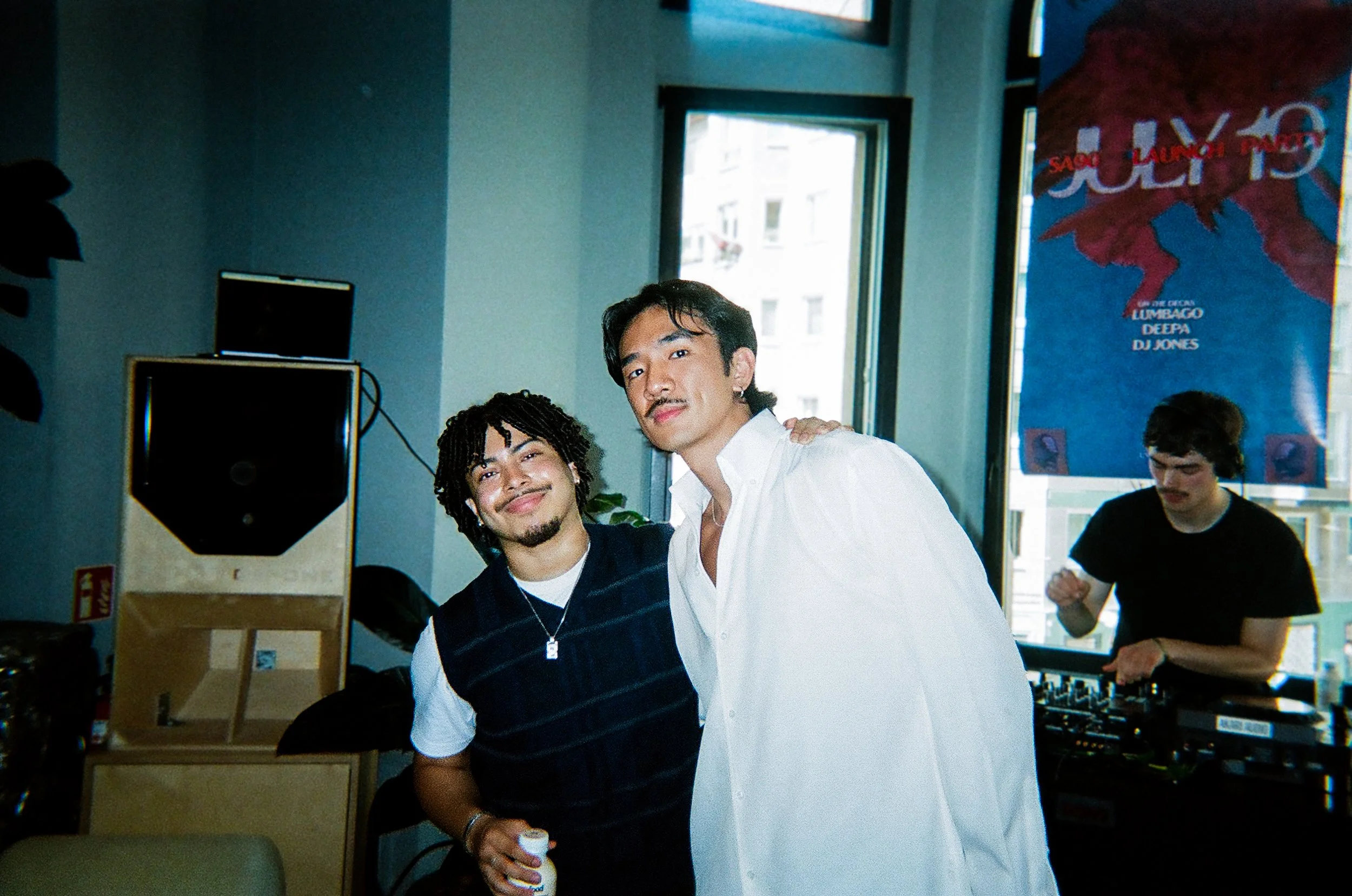 Two young men smiling and posing indoors, with one wearing a dark vest over a white T-shirt and the other in a white shirt, while a DJ plays music in the background.