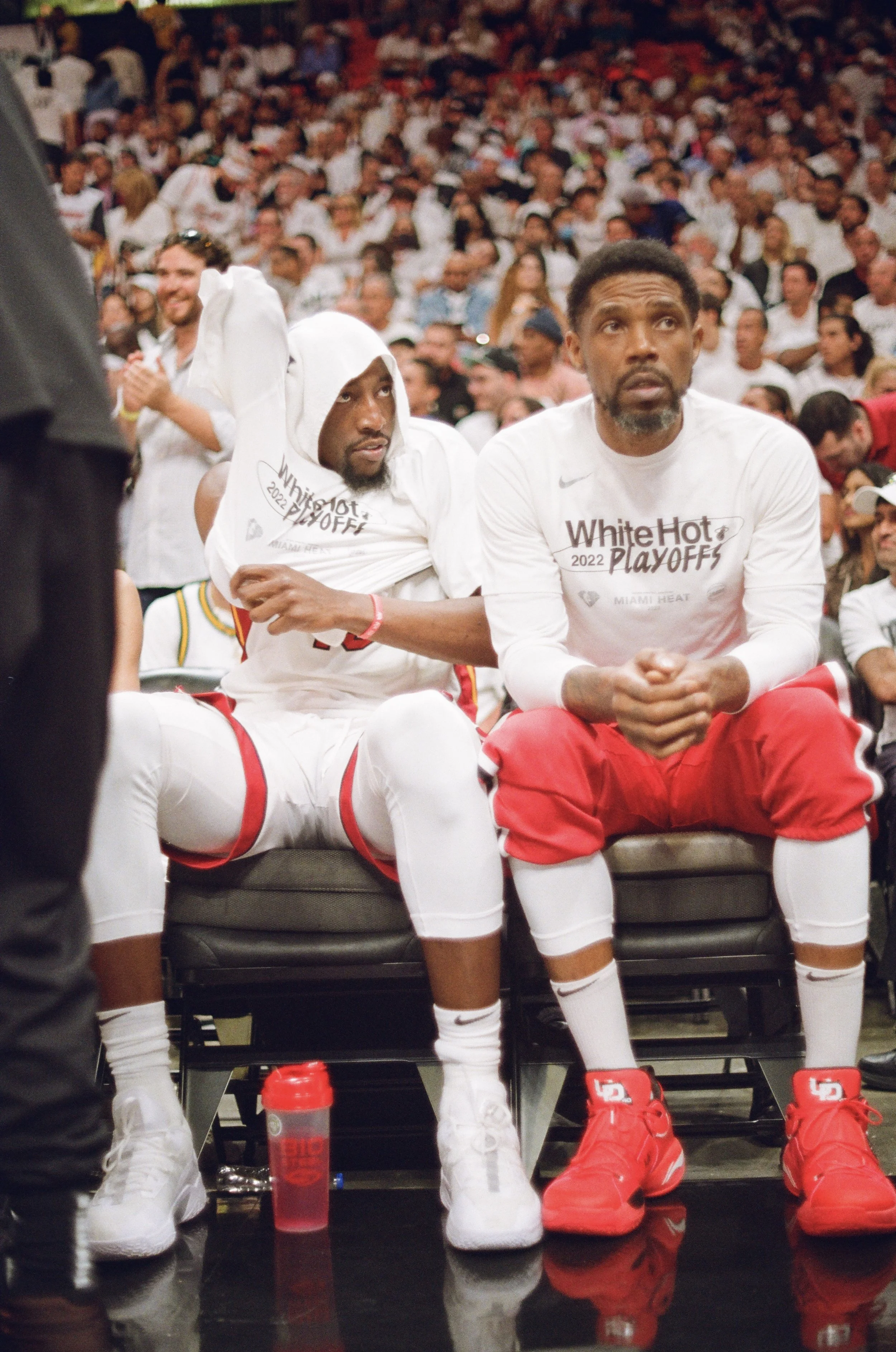 Two basketball players sitting on a bench during a game or event, wearing white uniforms with 'White Hot Playoffs' printed on them, surrounded by a crowd of spectators.