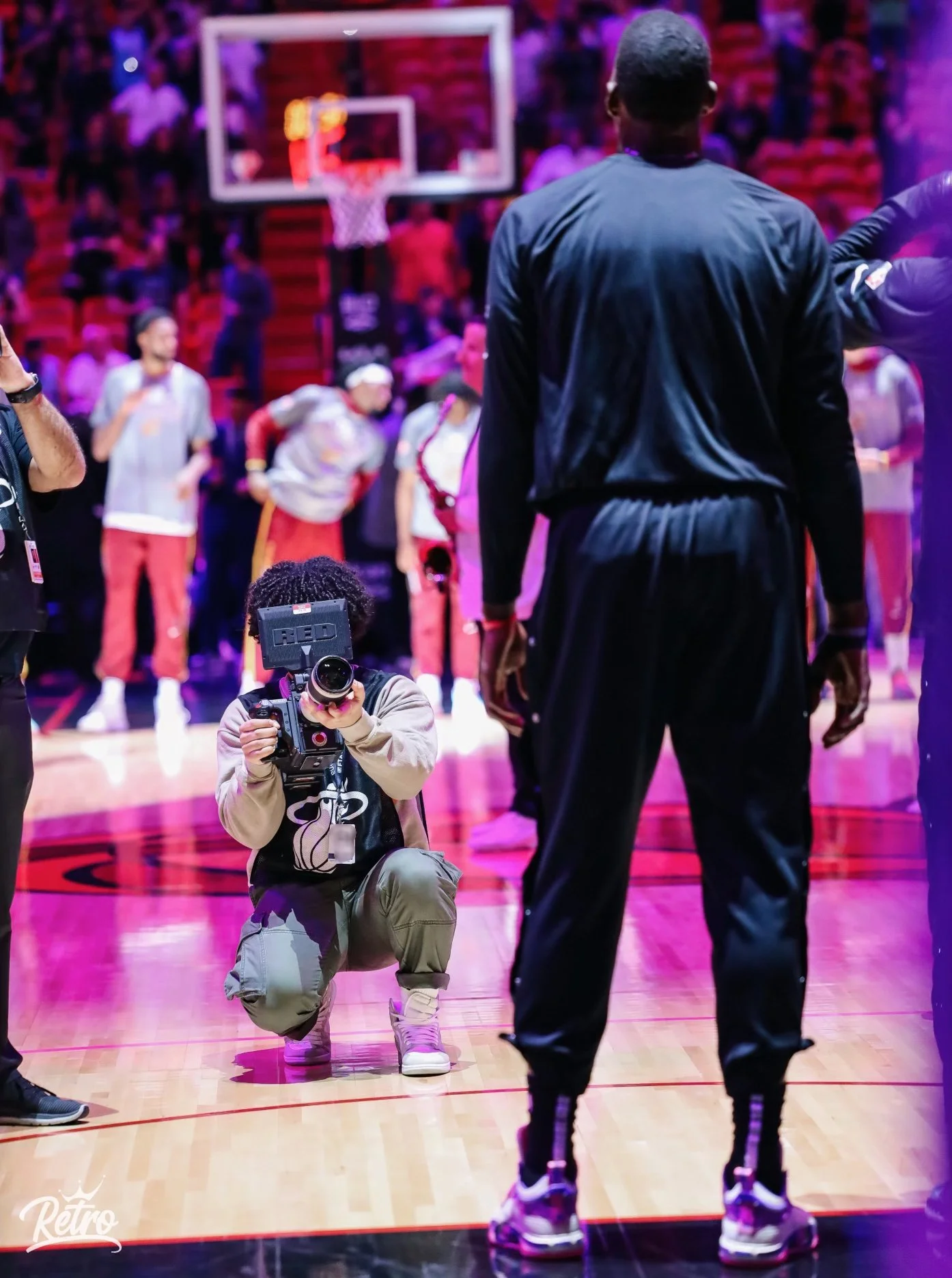 A photographer kneeling on a basketball court taking photos of a basketball game, with players and spectators in the background.