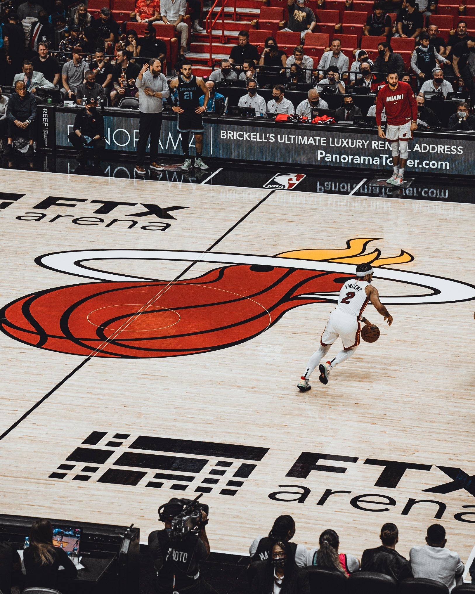 A basketball game at FTX Arena featuring a player from the Miami Heat dribbling the ball on the court with the Miami Heat logo, while players and coaches, some wearing masks, sit on the sidelines and a crowd watches from the stands.