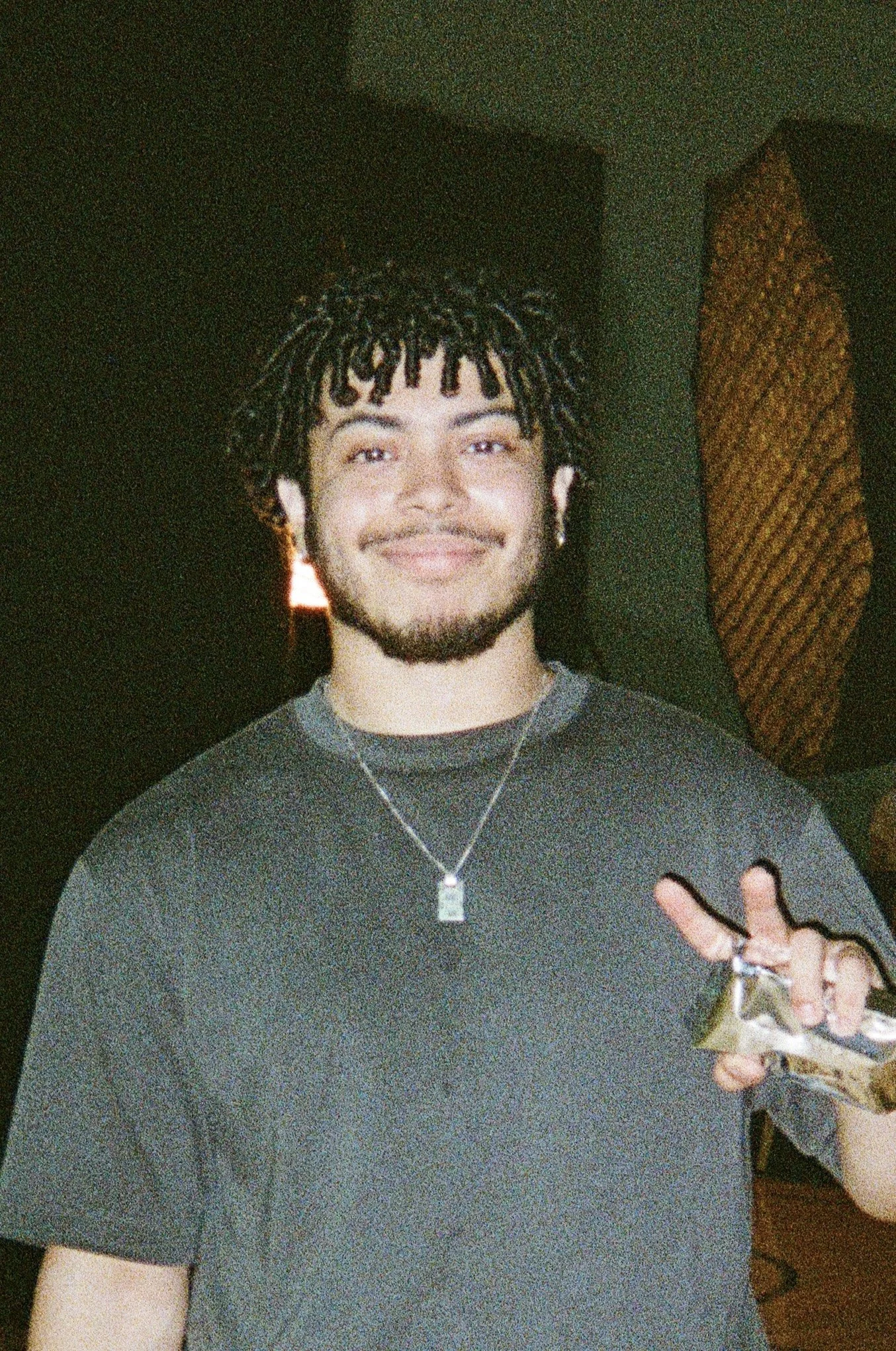 Young man with dark curly hair, beard, and earrings smiling, holding money, in a dimly lit room with decorative element in the background.