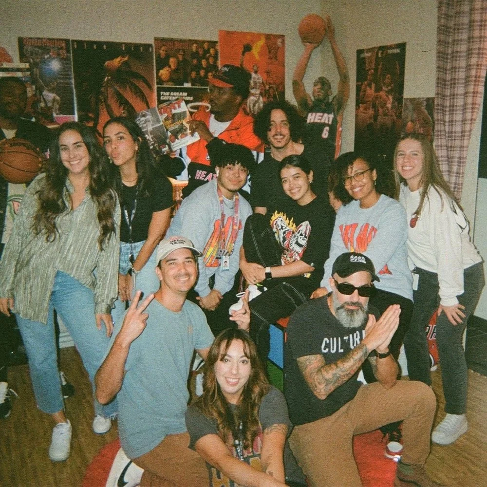 Group of people gathered in a room decorated with basketball posters, with some members wearing sports jerseys, posing for a photo.