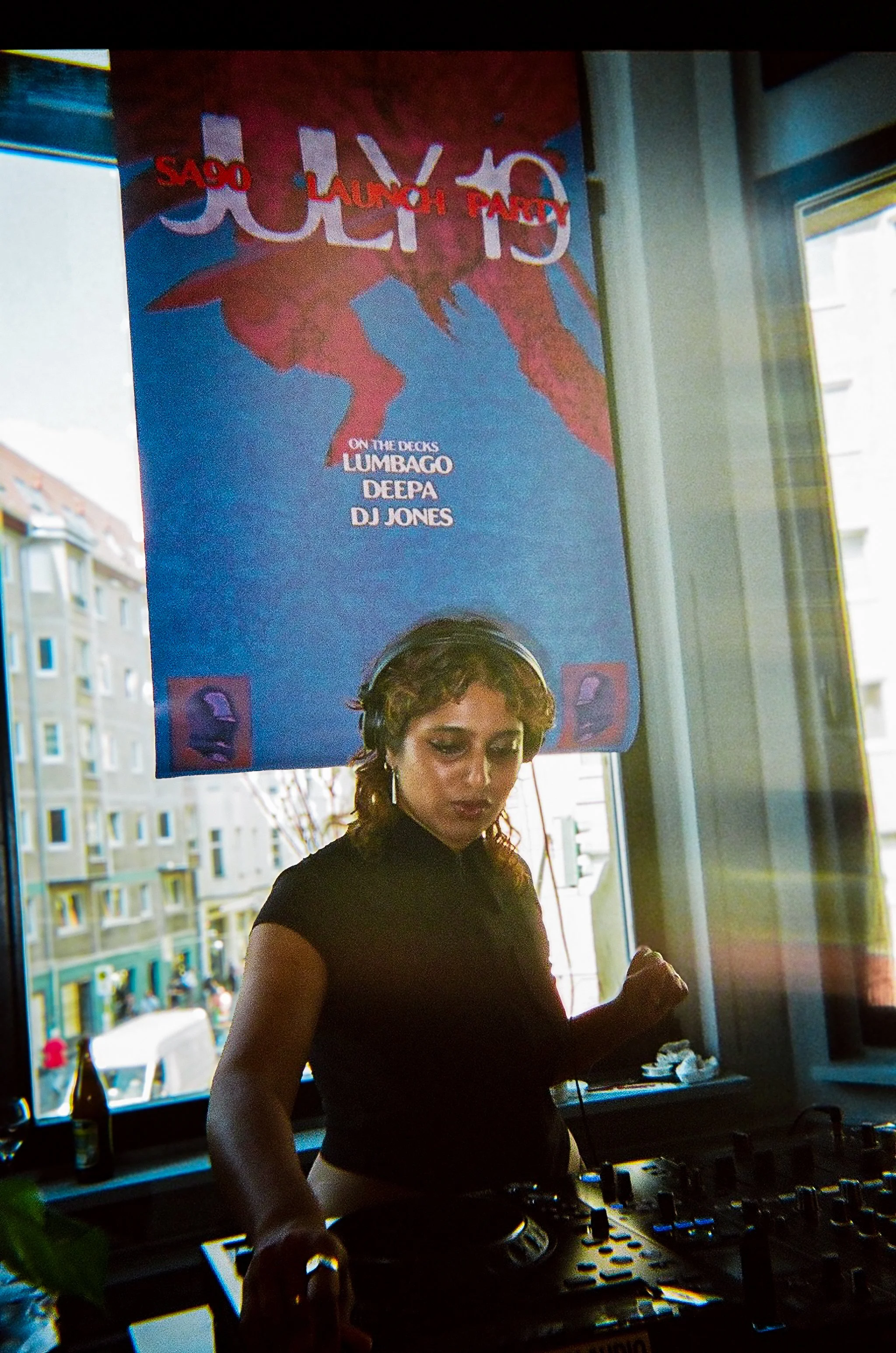 A female DJ is playing music with a DJ mixer in front of her, inside a room with large windows. Behind her, a colorful poster advertising a music event is visible, featuring the names 'LUMBAGO,' 'DEEPA,' and 'DJ JONES,' and a stylized graphic design.