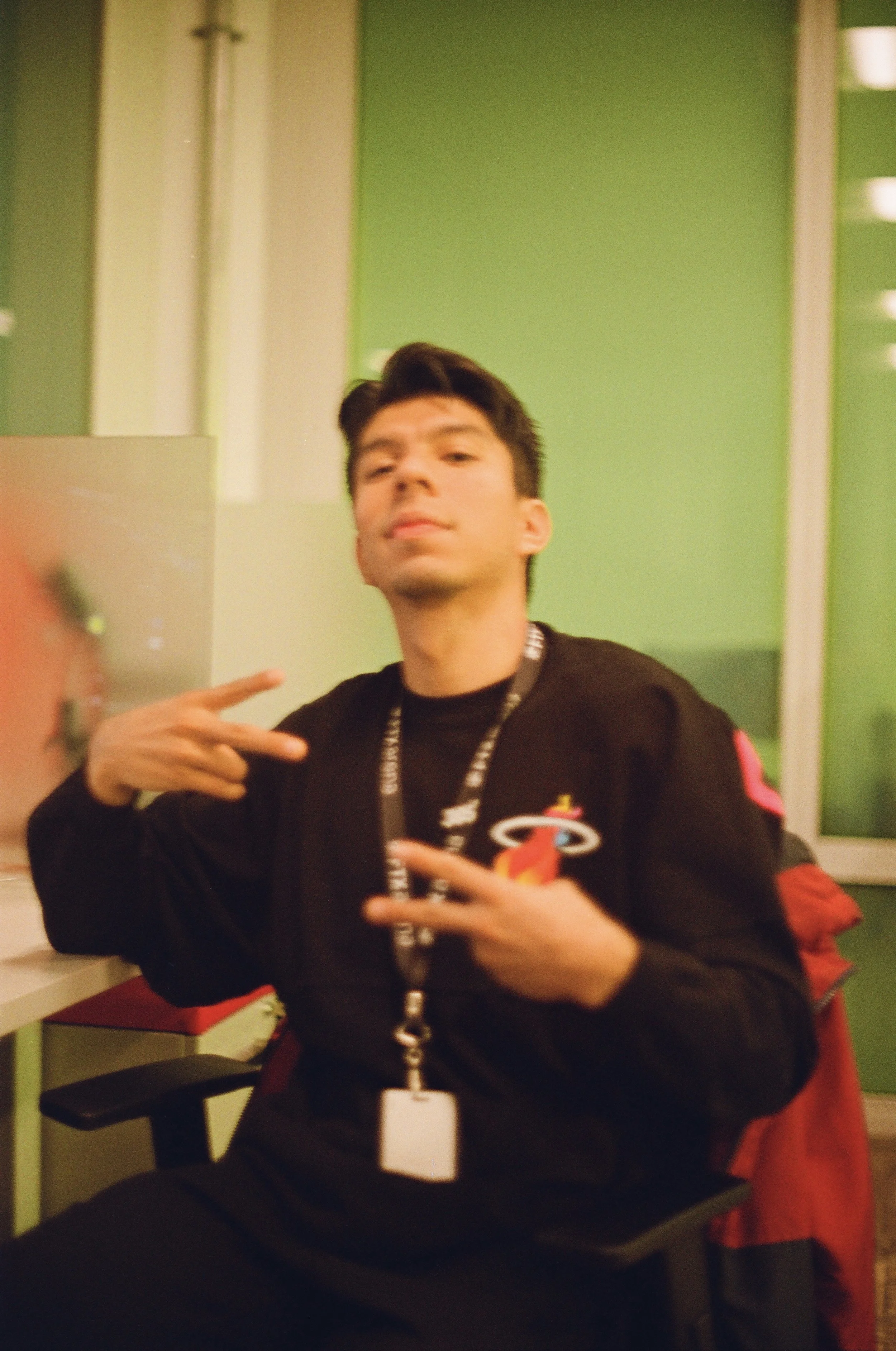 Young man with dark hair making a peace sign gesture with both hands, wearing a black sweatshirt and sitting at a desk in an office with green walls.