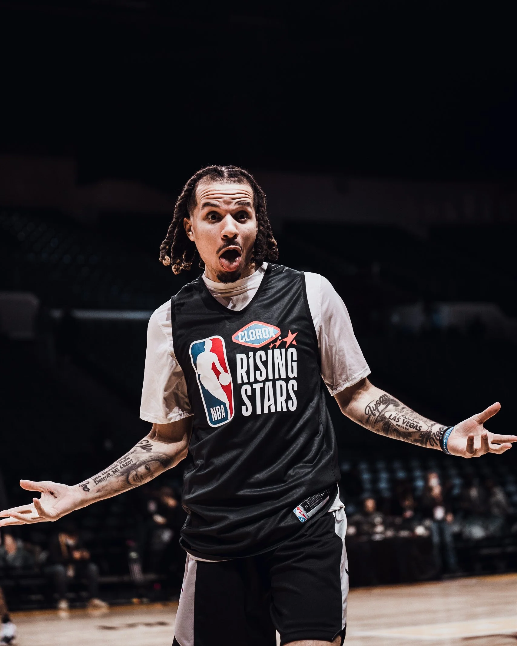 A basketball player with tattoos, wearing a black NBA Rising Stars jersey, making a surprised expression on the basketball court during a game.