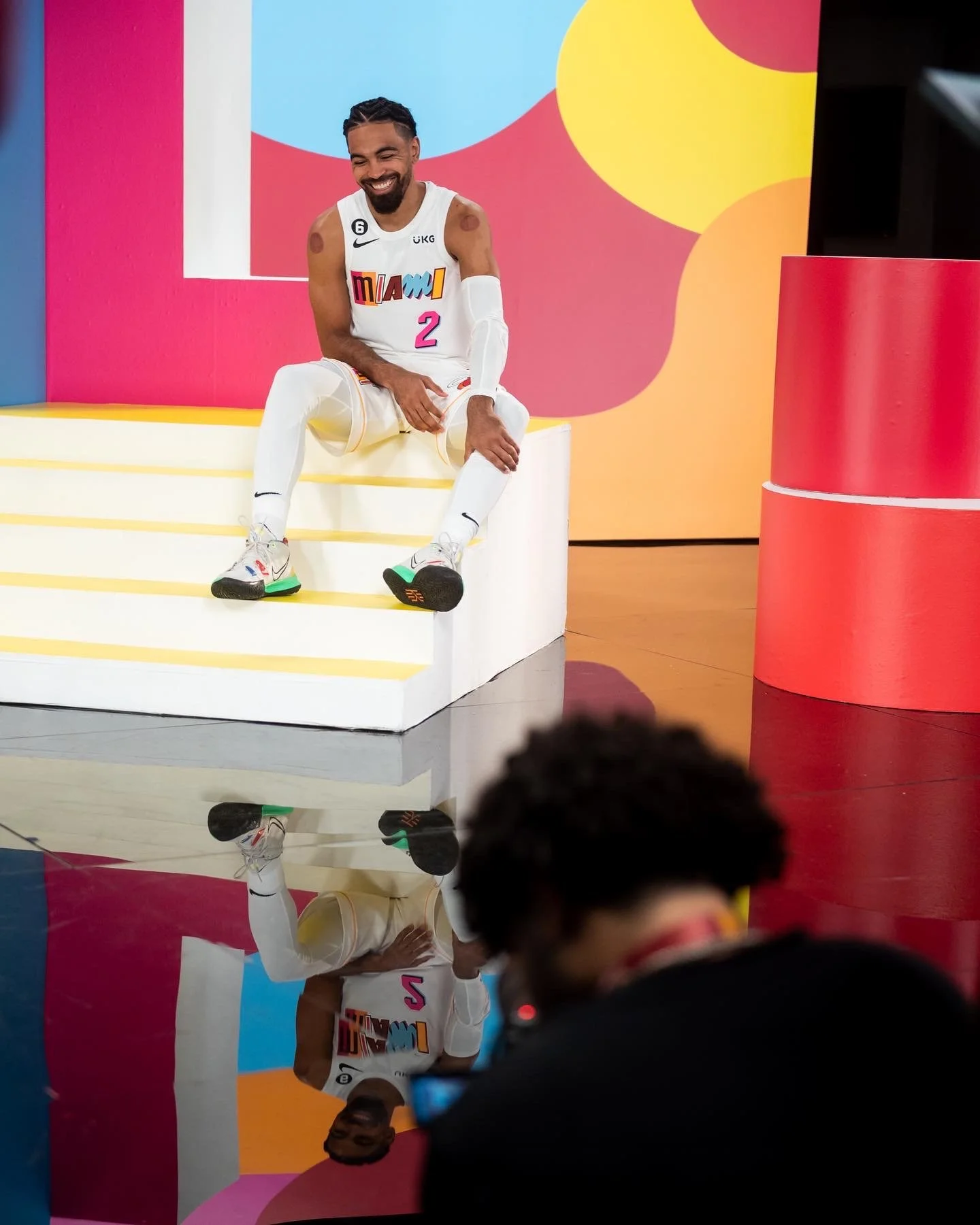 A smiling male athlete in a Miami Heat basketball uniform with the number 2, sitting on a yellow and white elevated platform with a colorful background, photocaptured by a photographer in the foreground.