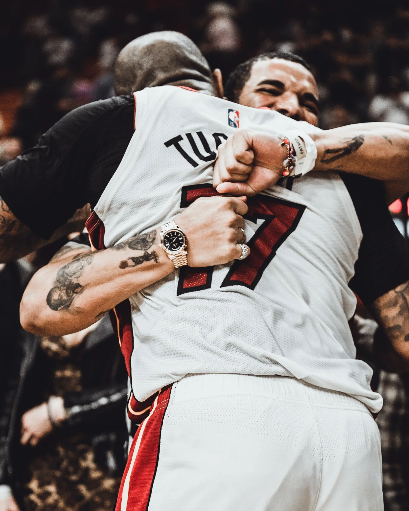 Two basketball players hugging in celebration, one wearing a Cleveland Cavaliers jersey with the name 'Tucker' and number 17, tattoos on both arms, a watch, and multiple rings, inside a crowded gym or arena.