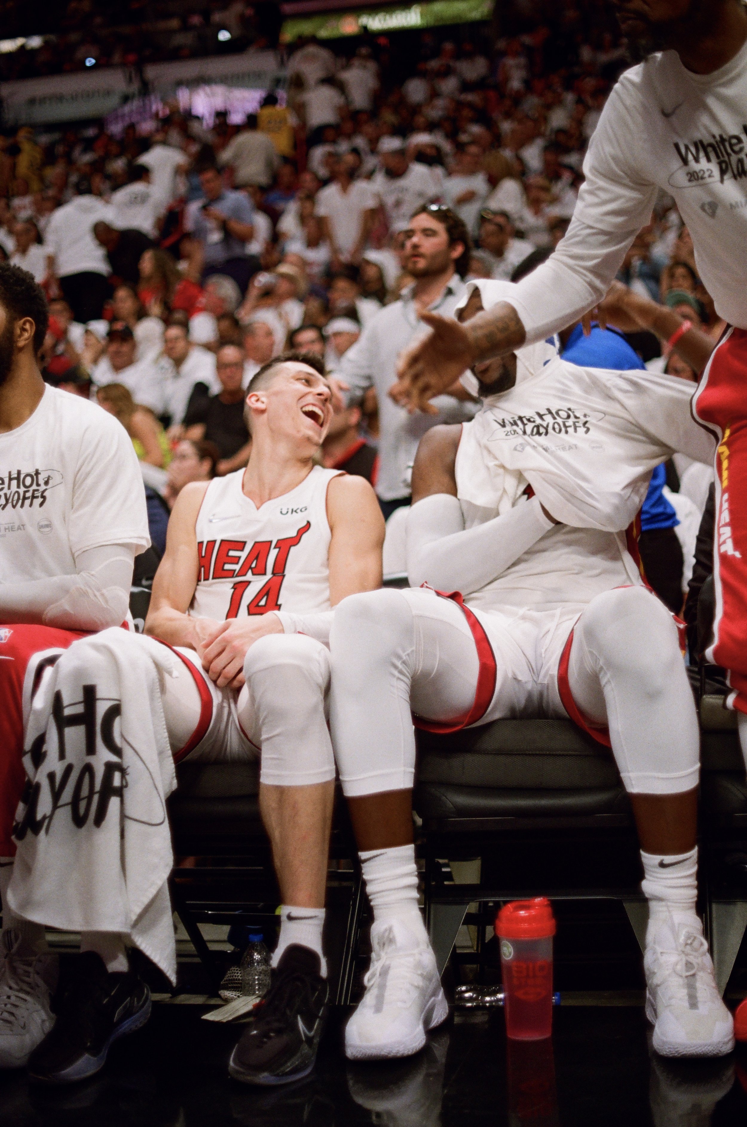 Miami Heat basketball players celebrating on the bench, with smiling and laughing faces, in an arena filled with fans.
