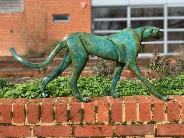 Bronze sculpture of a walking panther or similar big cat animal placed on a brick ledge in front of a building with windows and plants.