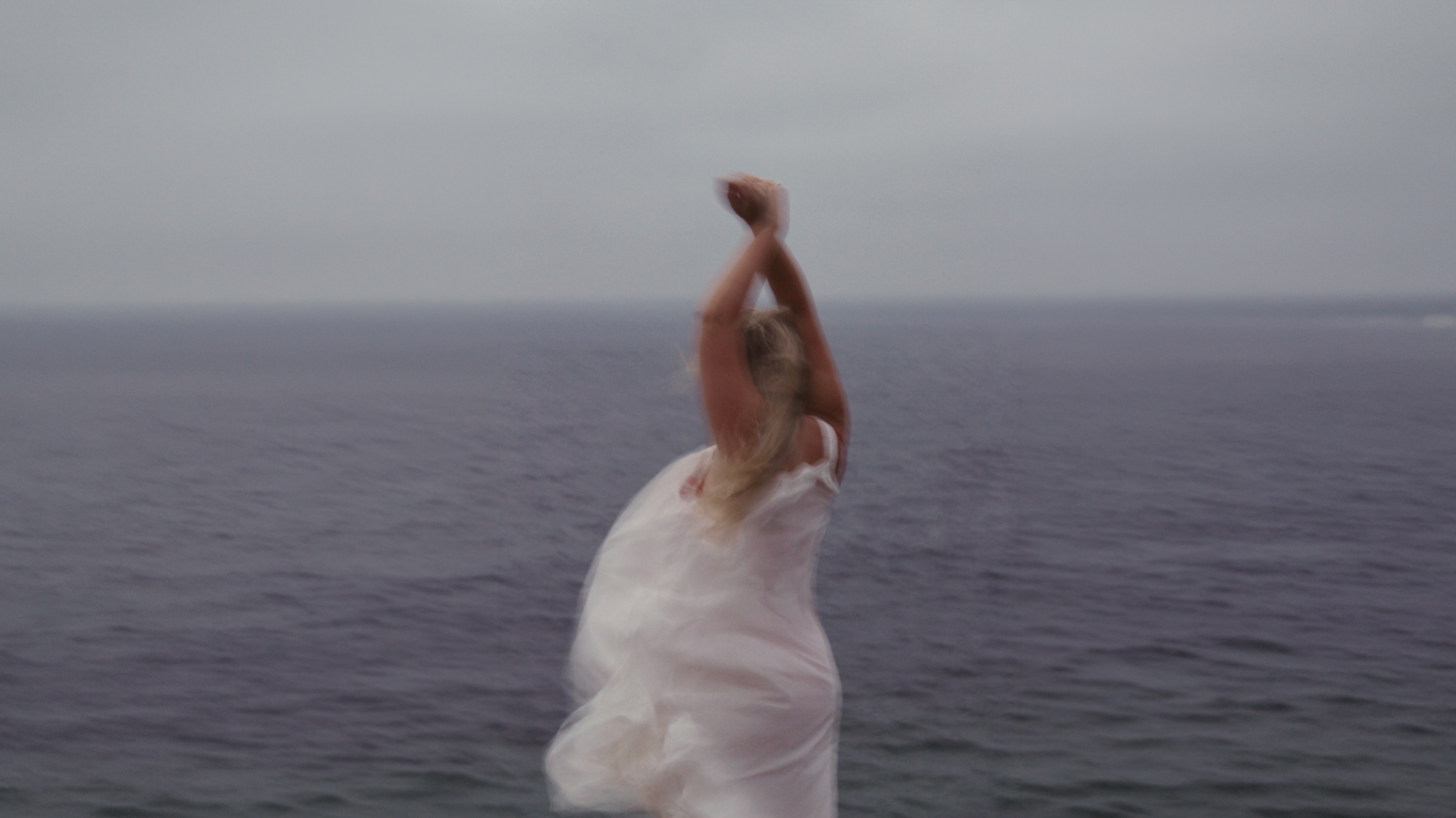 A woman in a white dress standing on an ocean shoreline, with arms raised and crossed above her head, blurred motion, cloudy sky in background.