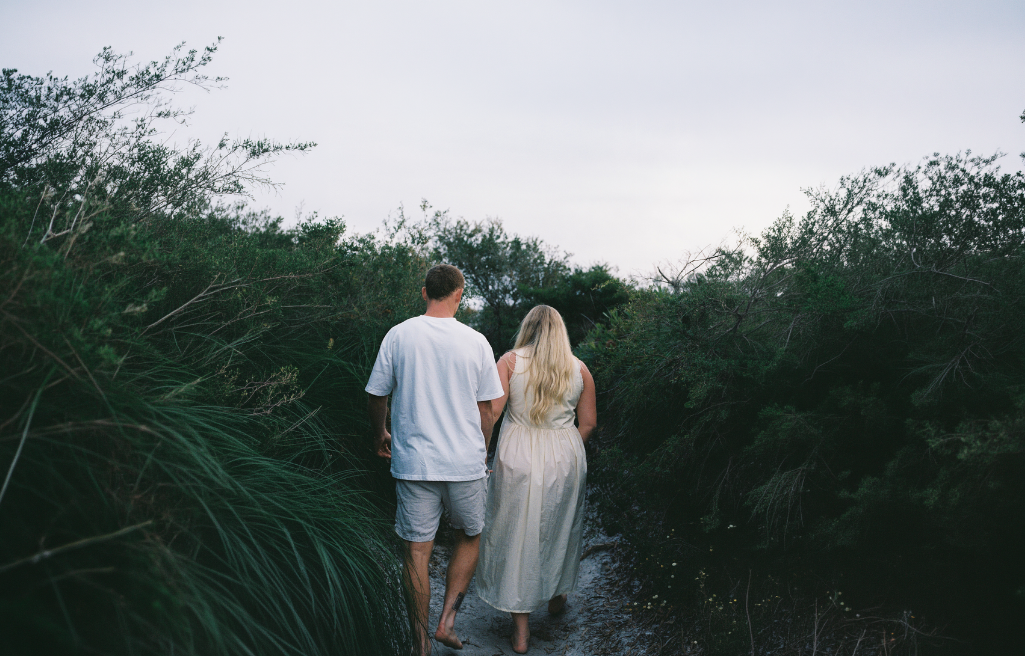 A man and woman walking hand in hand through a narrow stream surrounded by greenery at dusk.