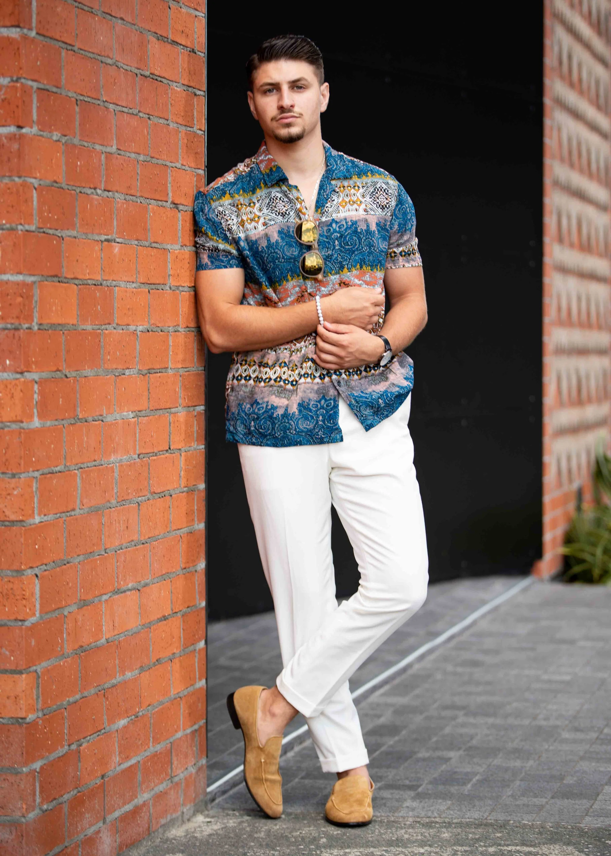 A young man standing against a brick wall, wearing a patterned short-sleeve shirt, white pants, and tan shoes, with sunglasses hanging on his shirt, and a watch and bracelet on his left wrist.