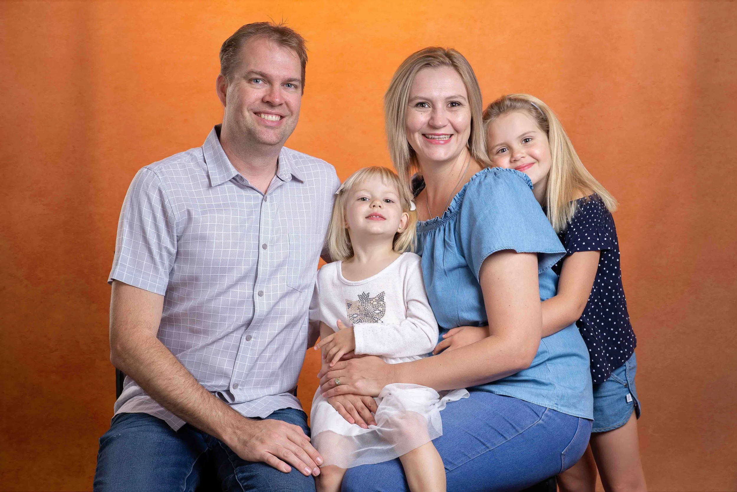 A smiling family of four, including a father, mother, and two young daughters, posing together against an orange backdrop.