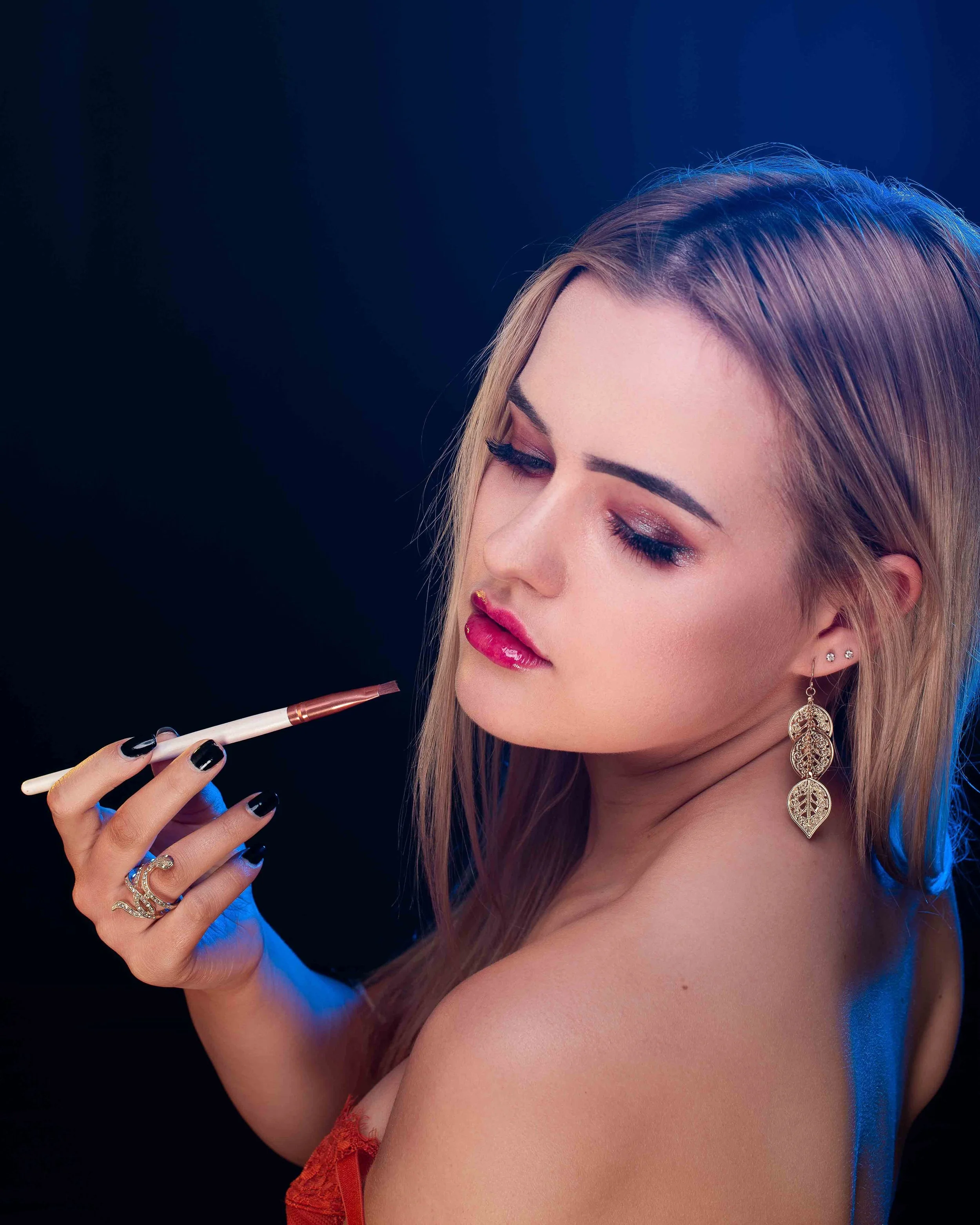 A woman with long light brown hair, wearing makeup and gold earrings, holding a makeup brush near her face against a dark background.