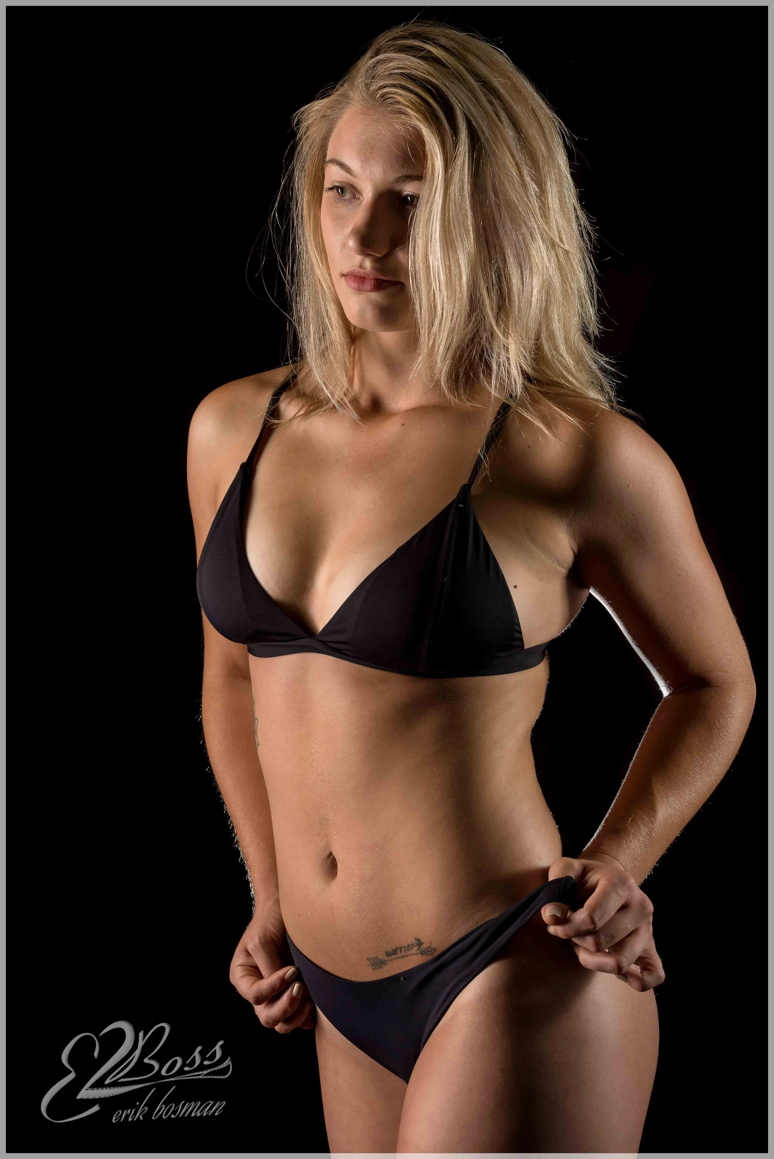 A woman with blonde hair in black lingerie standing against a black background with soft lighting.