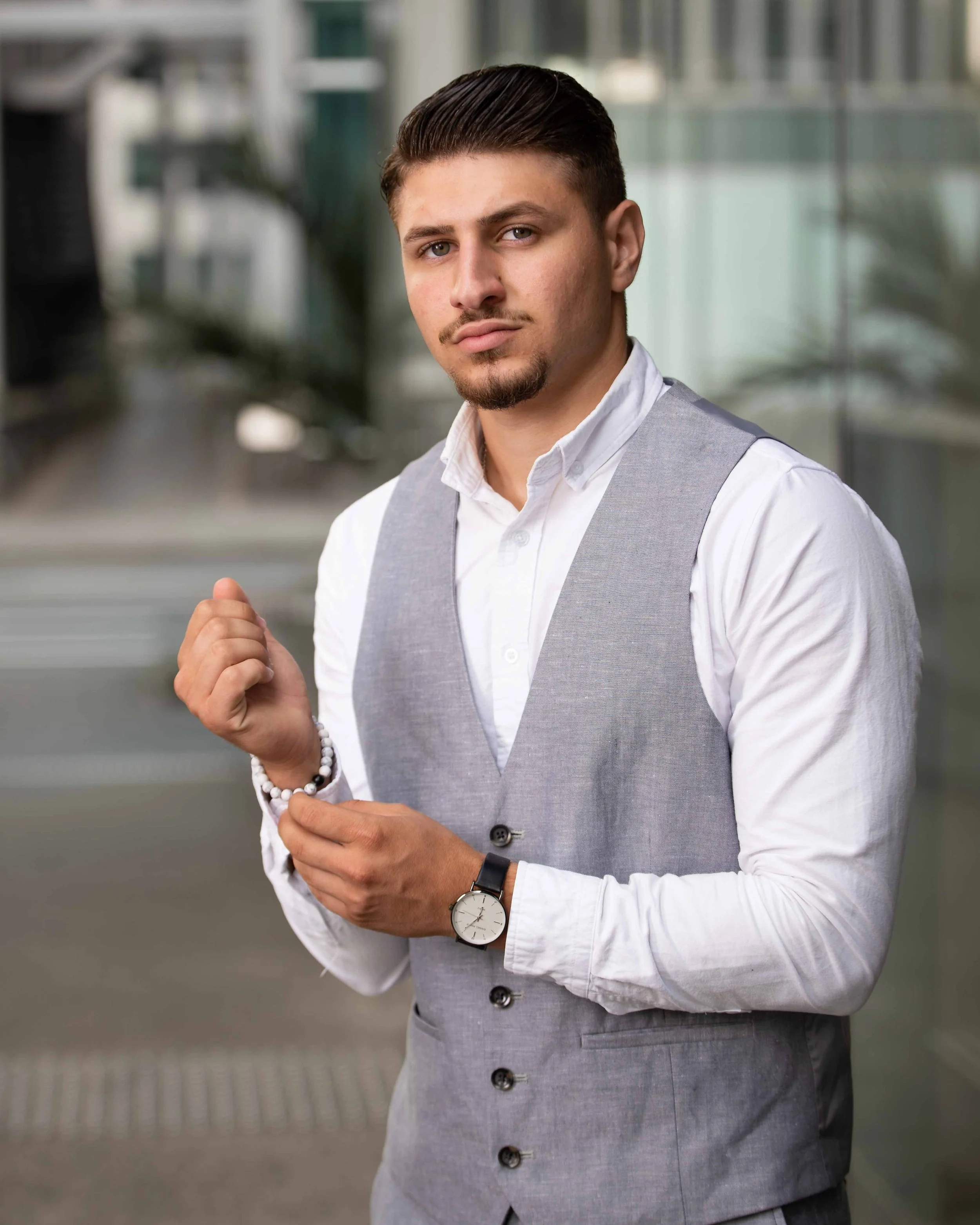 A young man in a white shirt and gray vest standing outside a modern building, adjusting his wristwatch and holding a beaded bracelet.