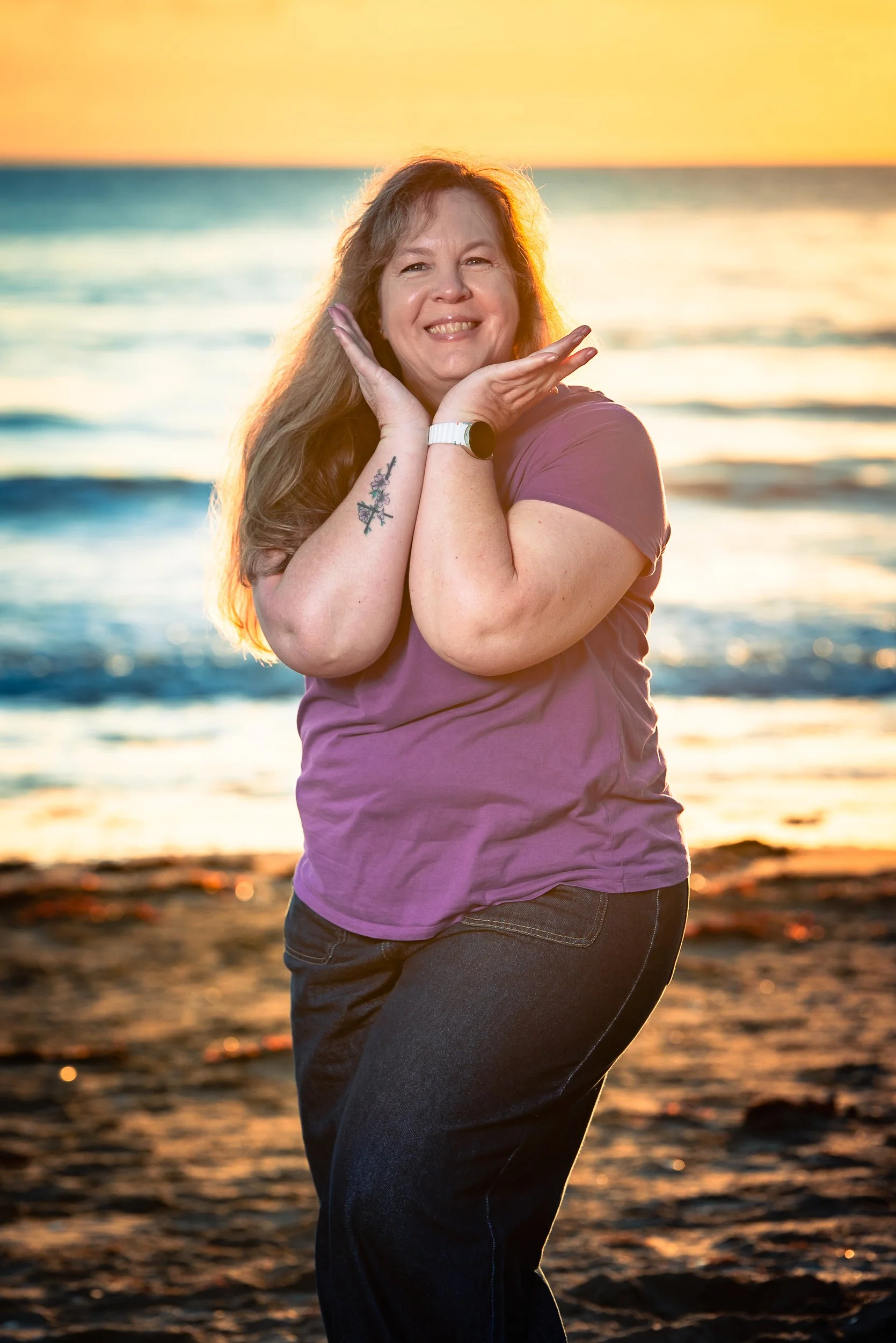 A woman smiling at the camera poses on a beach at sunset, with her hands framing her face, wearing a purple t-shirt and jeans.