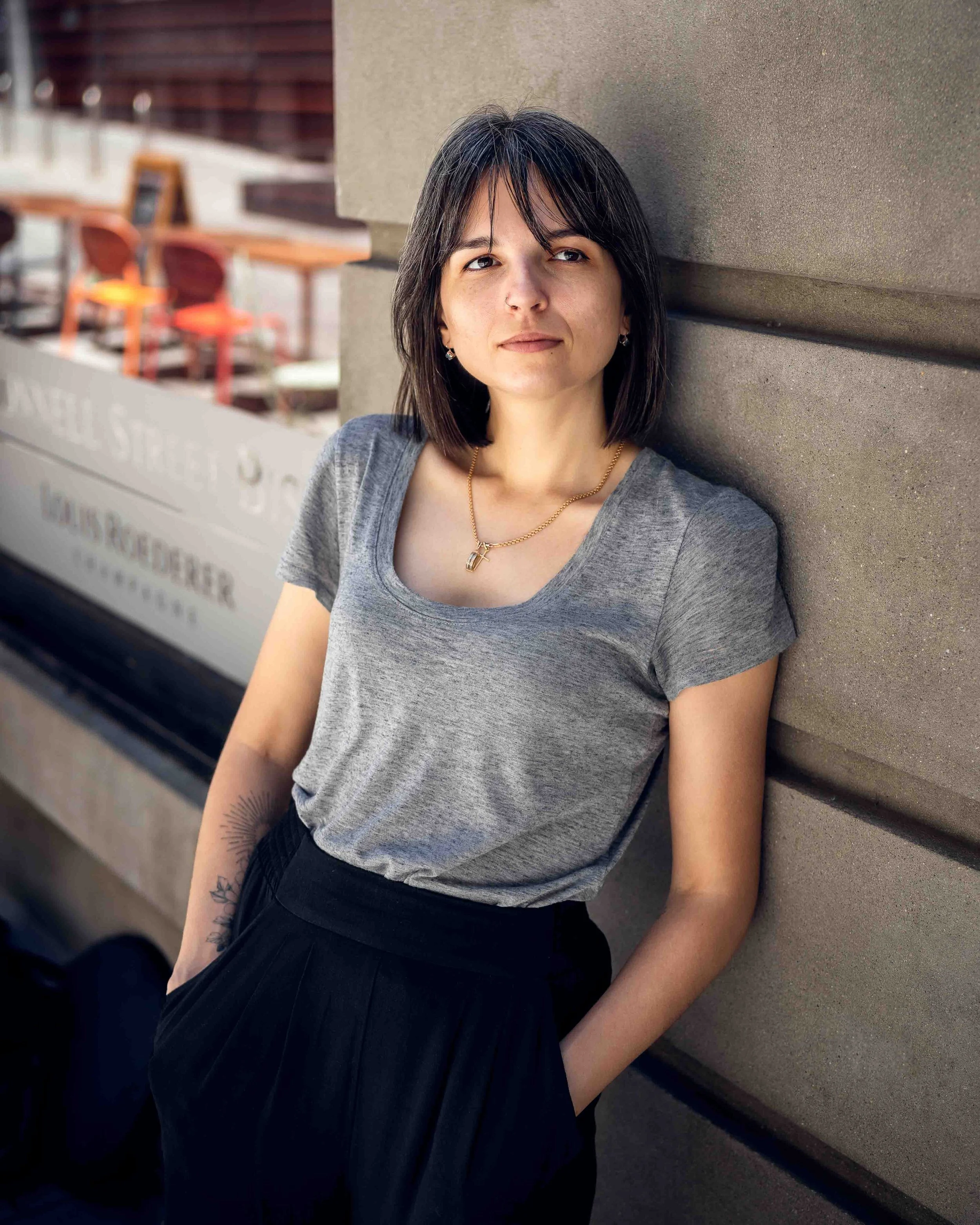 A woman with shoulder-length dark hair, wearing a gray t-shirt and black pants, leaning against a textured beige wall outdoors with her hands in her pockets, and looking off to the side.