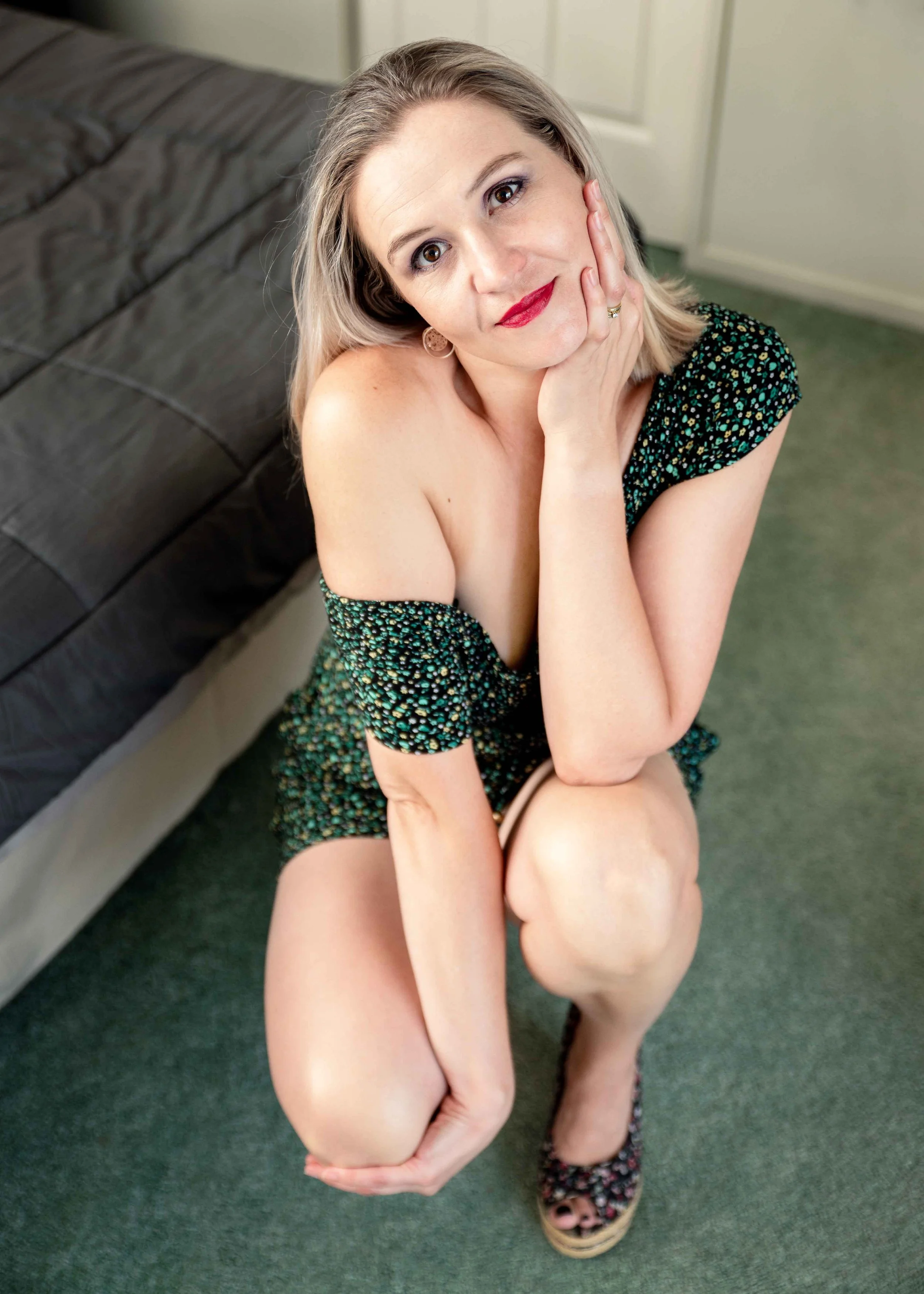 A woman sitting on the floor, wearing a patterned off-shoulder dress, with one hand resting on her cheek, looking at the camera, with a bed in the background.