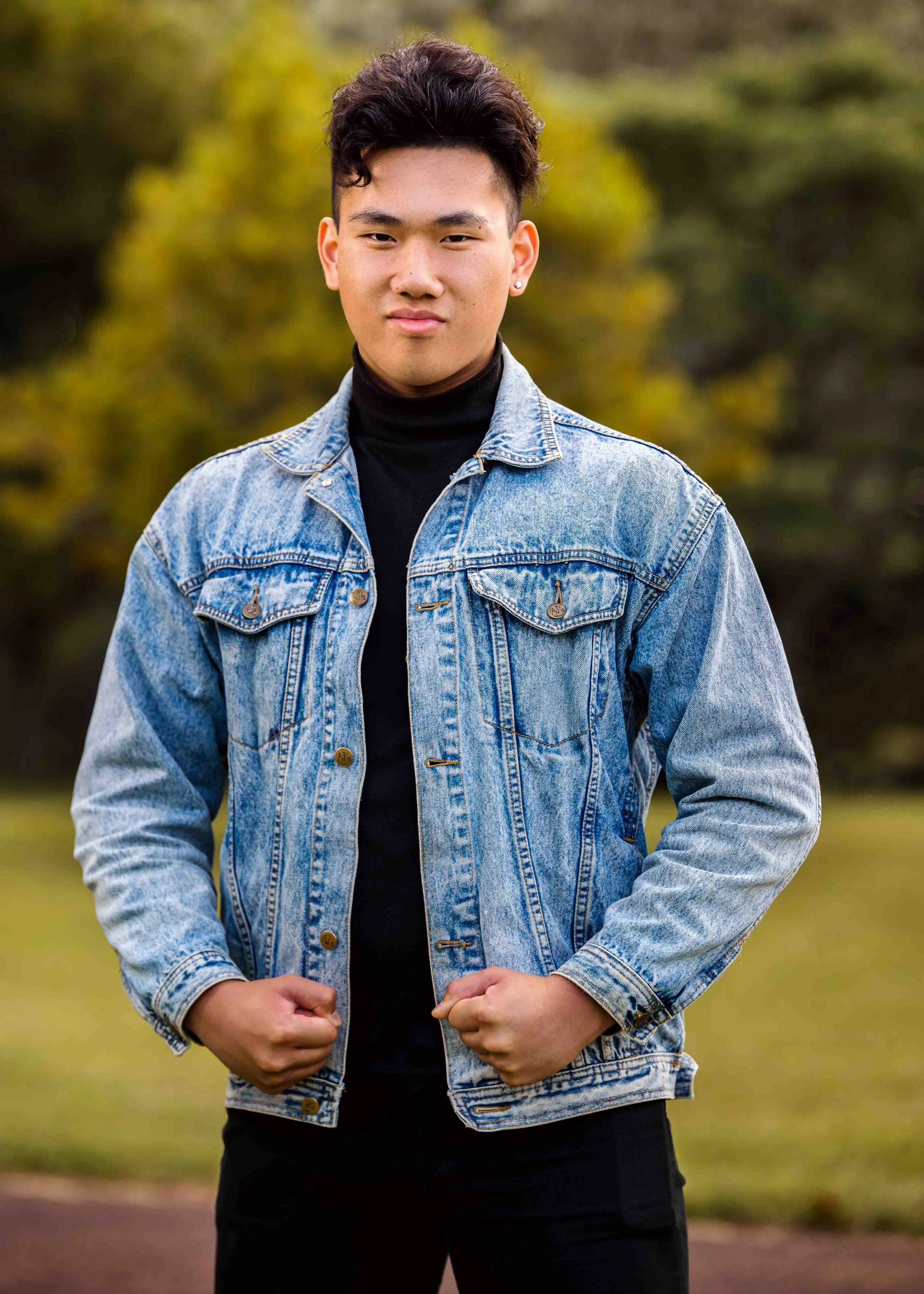 A young man standing outdoors in a park with trees in the background, wearing a denim jacket and a black turtleneck.