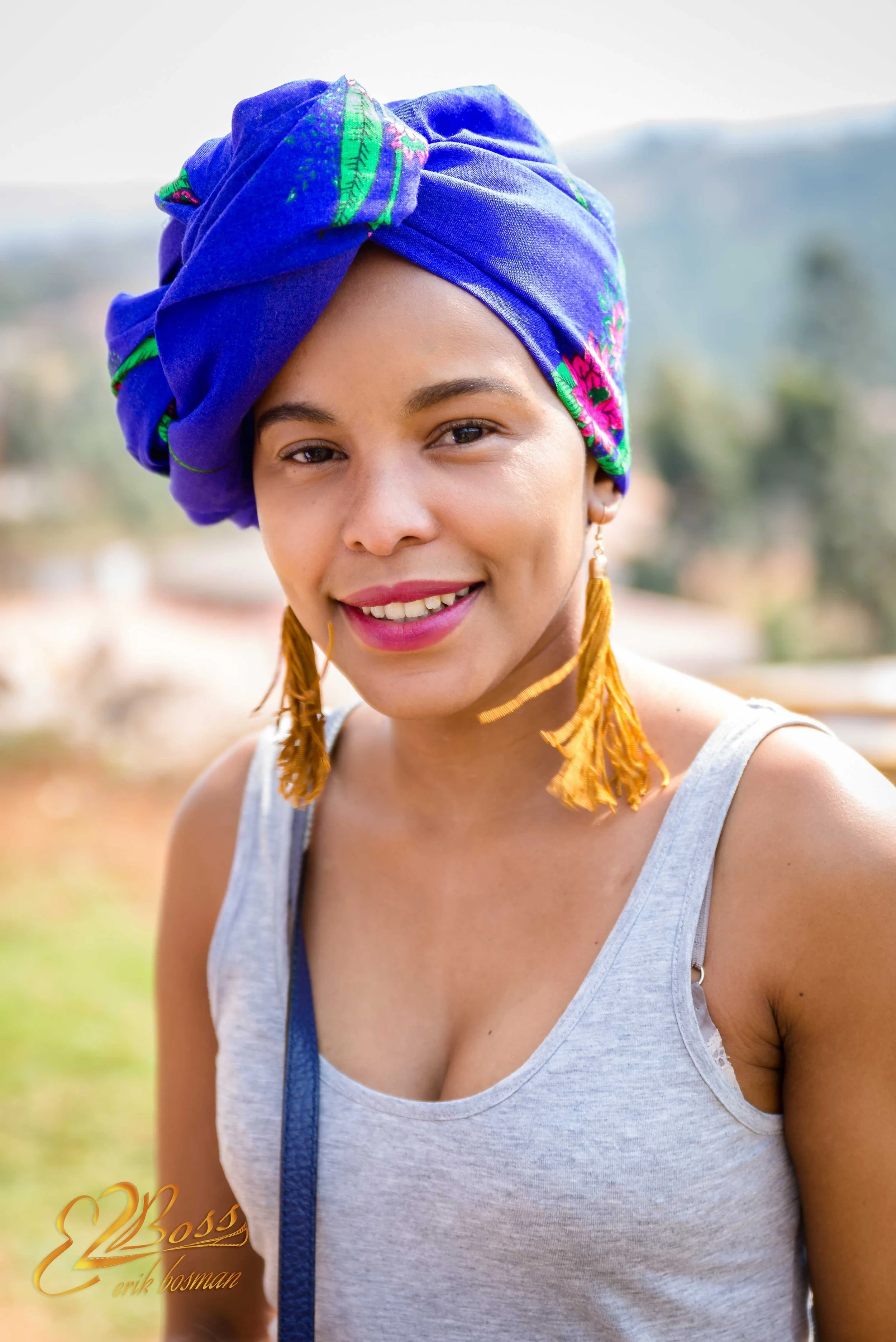 A woman outdoors wearing a vibrant blue headwrap with green and pink embroidery, gold tassel earrings, and a gray tank top, smiling at the camera.