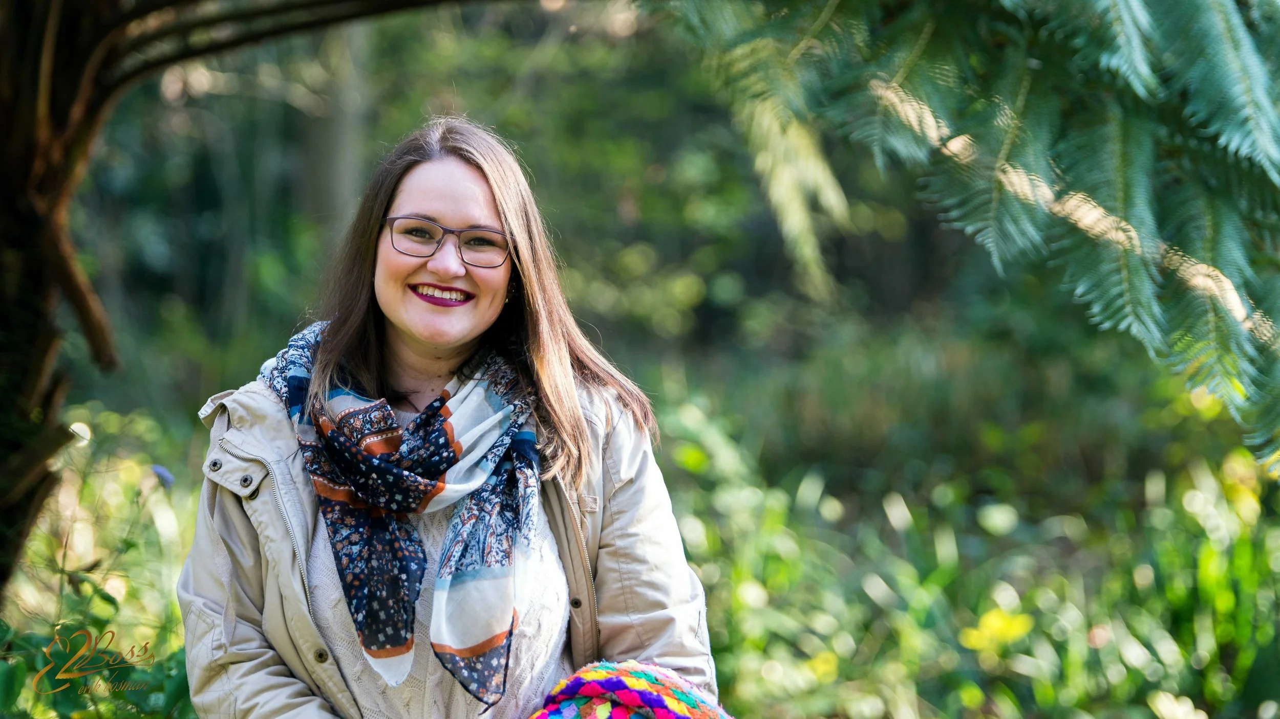 A smiling woman with glasses and a colorful scarf standing outdoors in a lush green forest.