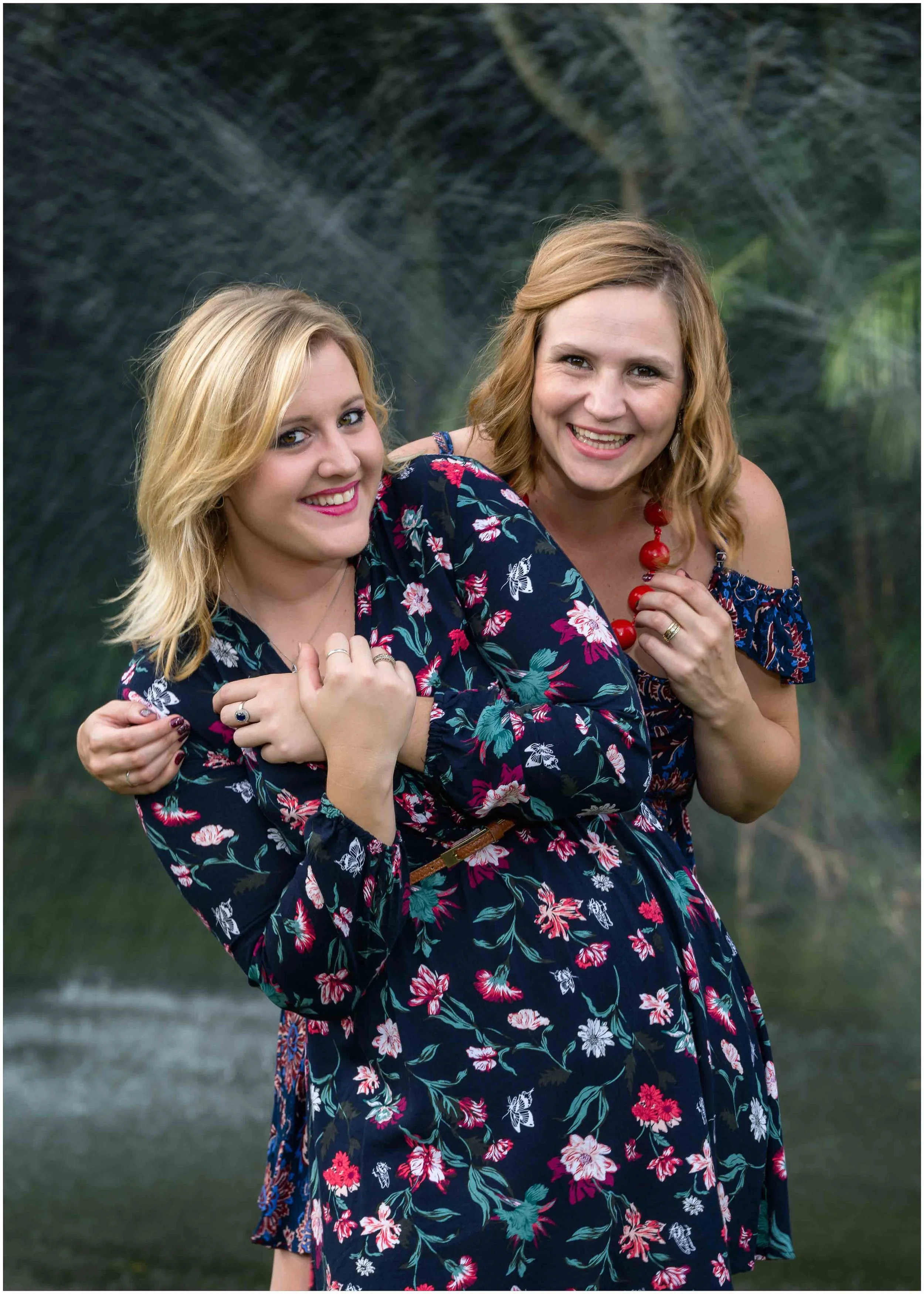 Two women in floral dresses smiling outdoors with trees and water in the background.