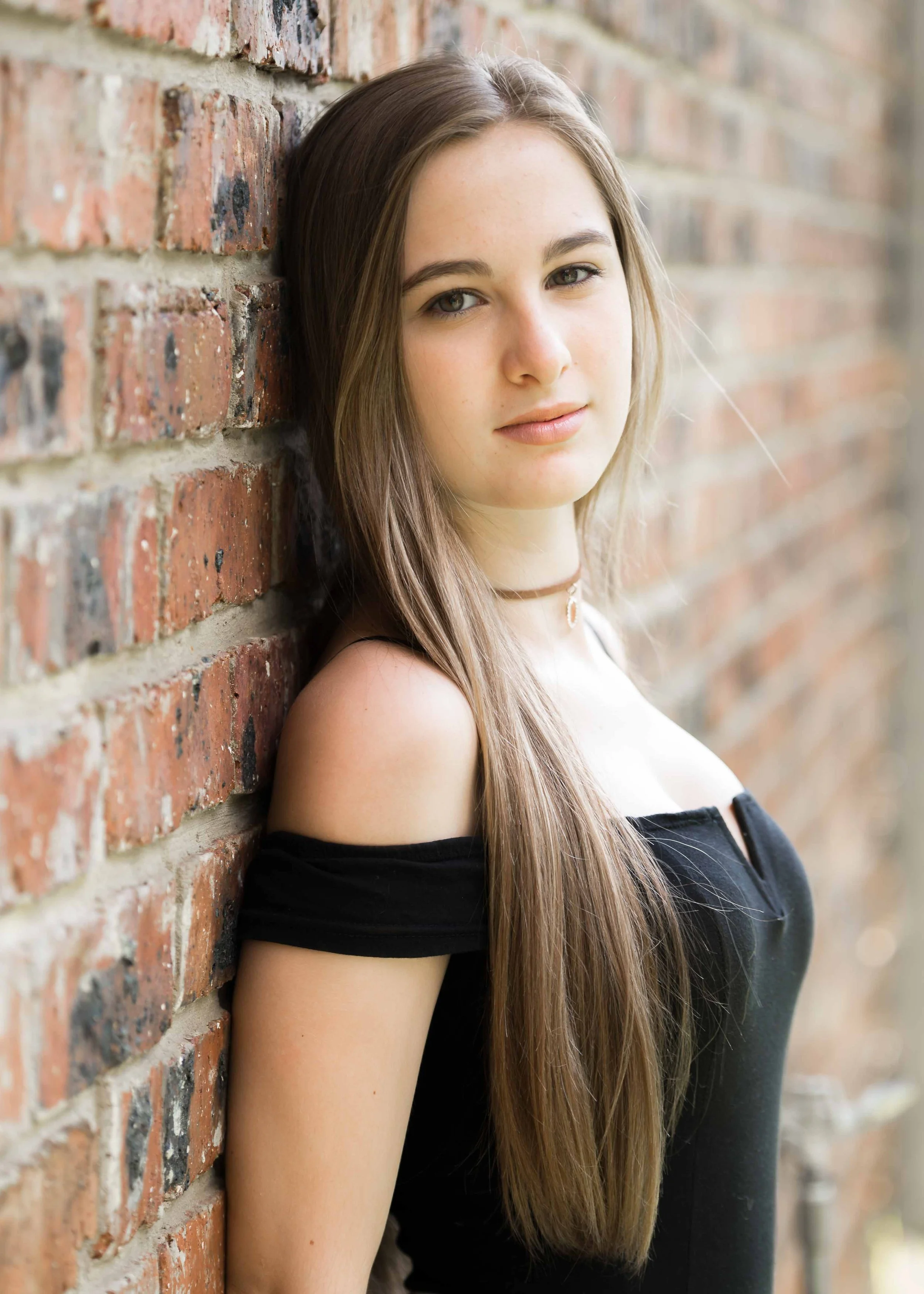 A young woman with long brown hair and fair skin leaning against a brick wall, wearing a black off-shoulder top and a choker necklace, looking at the camera.