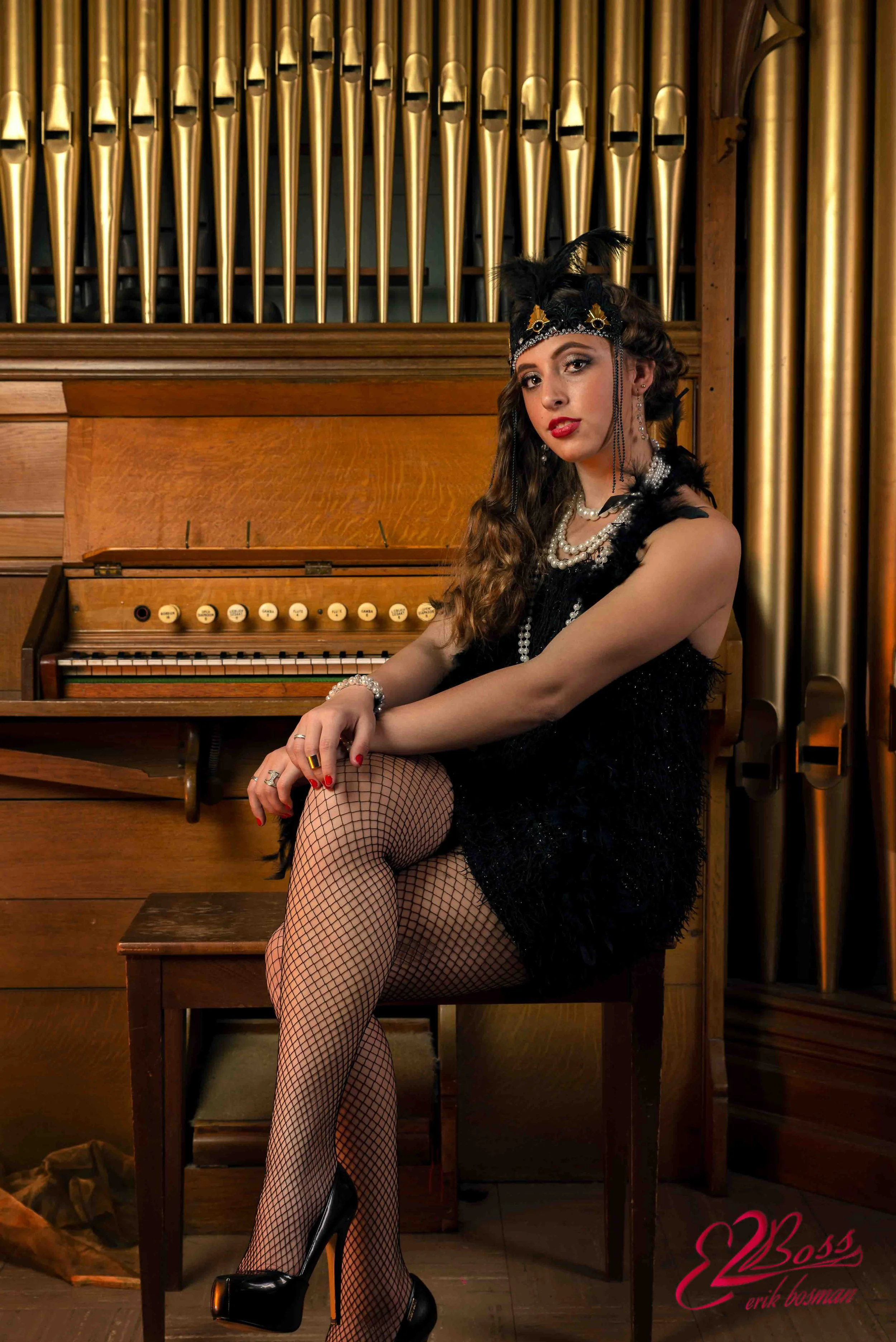 A woman dressed in a vintage 1920s flapper costume sitting on a wooden chair in front of an organ, with a backdrop of large, gold pipes. She is wearing a black feathered headband, black dress, pearl jewelry, fishnet stockings, and high heels.
