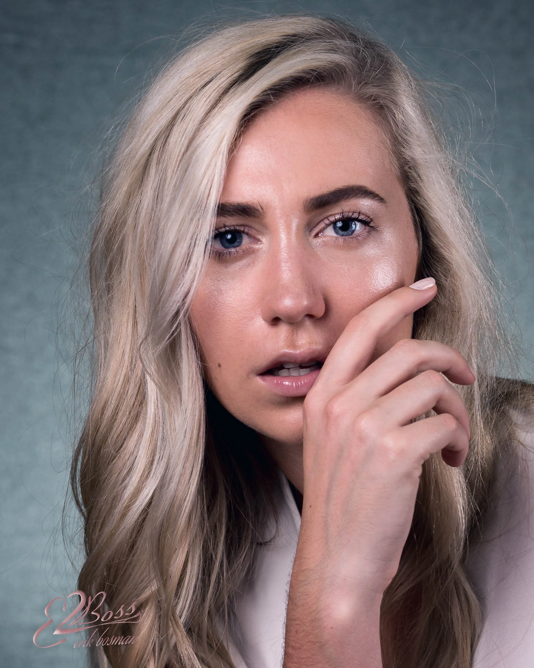 Close-up portrait of a young woman with long blonde hair and blue eyes, touching her face with one hand, against a gray background.