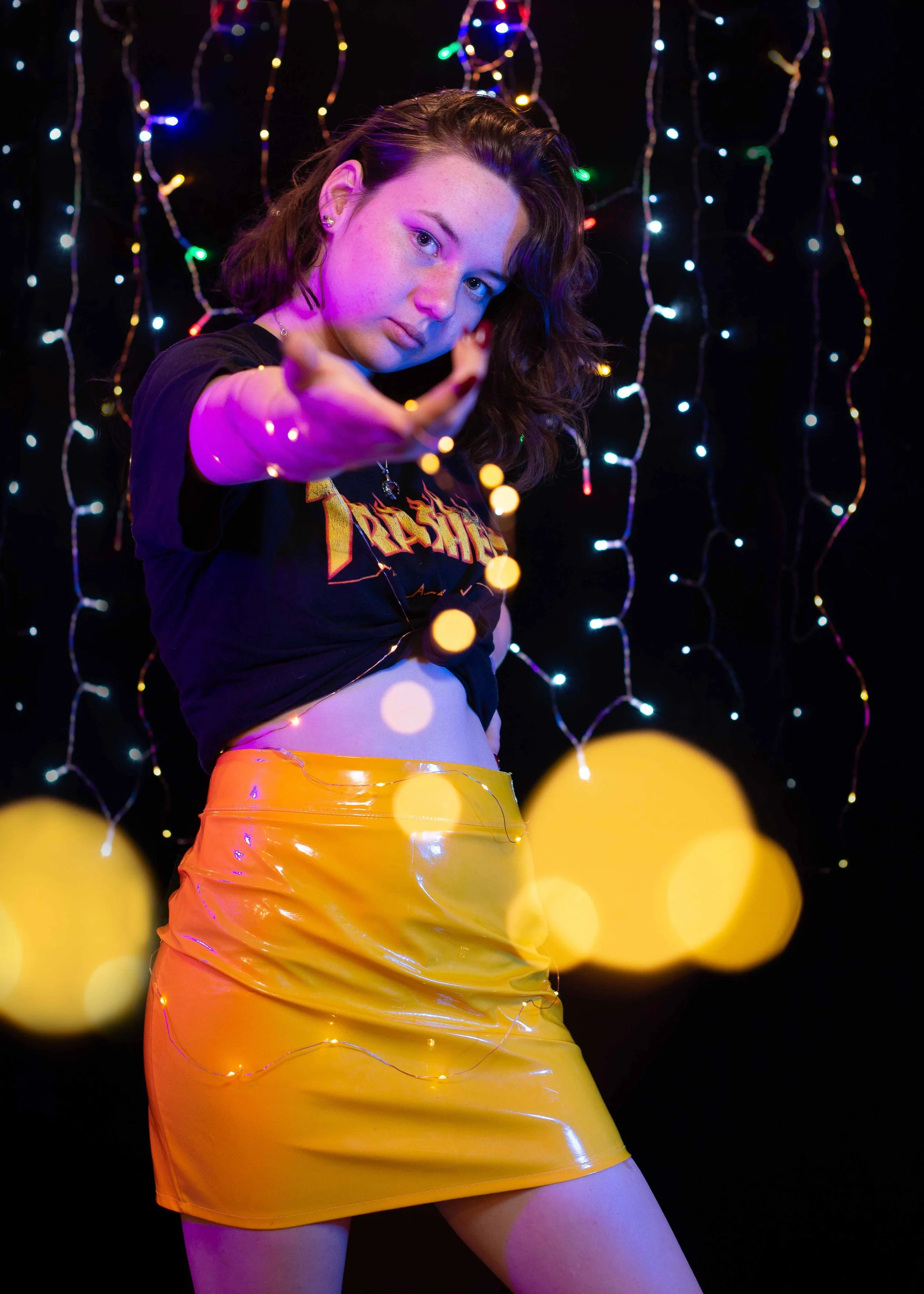 A young woman with shoulder-length brown hair poses confidently in front of a dark background decorated with colorful string lights. She wears a black T-shirt with a logo, a shiny yellow mini skirt, and is reaching toward the camera with her hand, cr