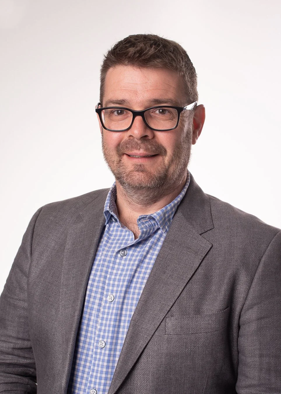 A close-up portrait of a man with short brown hair, glasses, and a beard wearing a gray blazer and a blue checkered shirt against a plain white background.