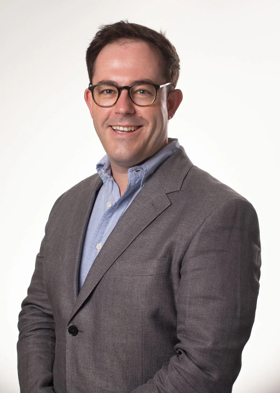 Portrait of a man with glasses in a suit jacket and light blue collared shirt smiling against a white background.