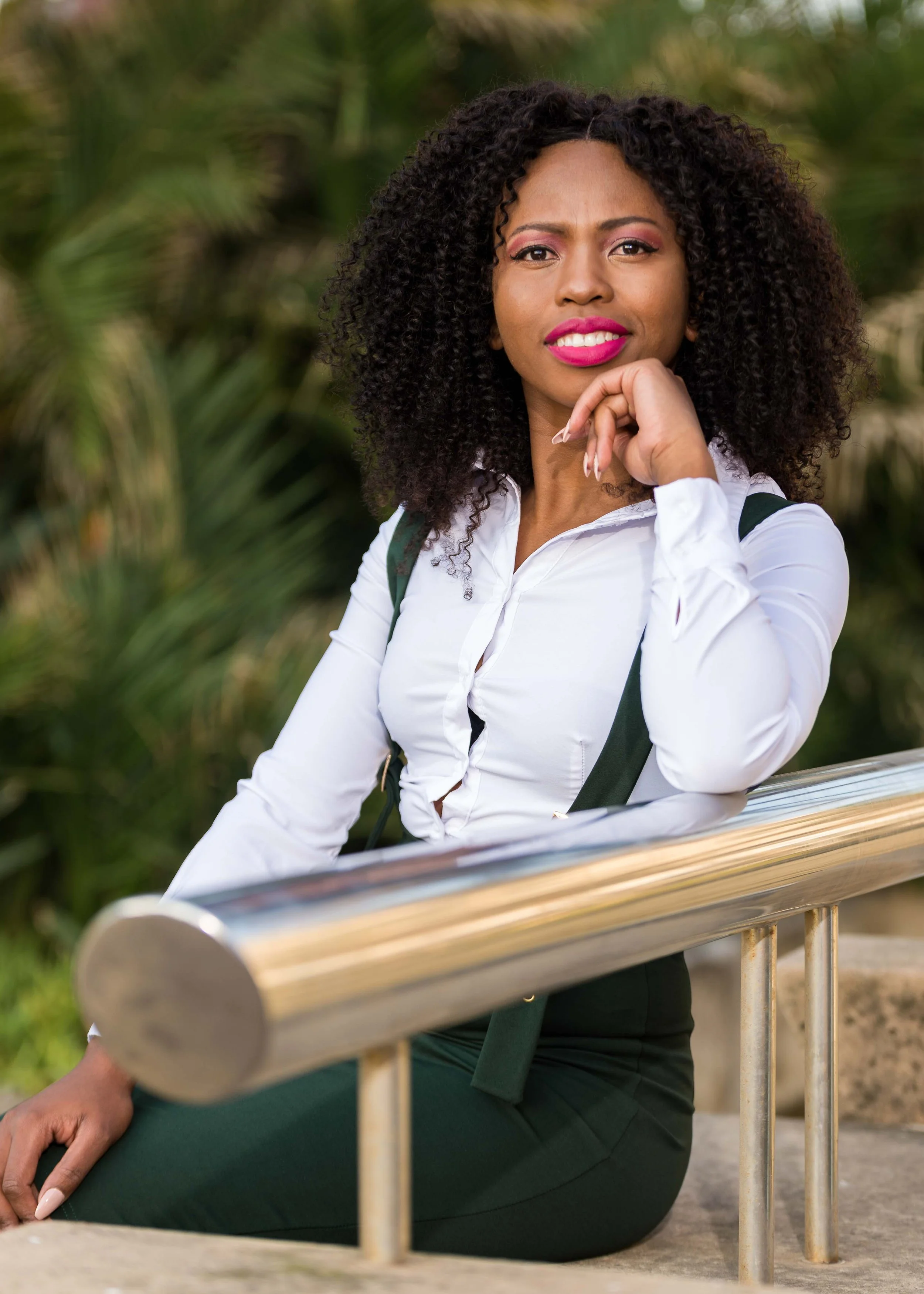 A woman with curly hair wearing a white blouse with suspenders, sitting on stairs outside, resting her arm on a metal railing, with greenery in the background.