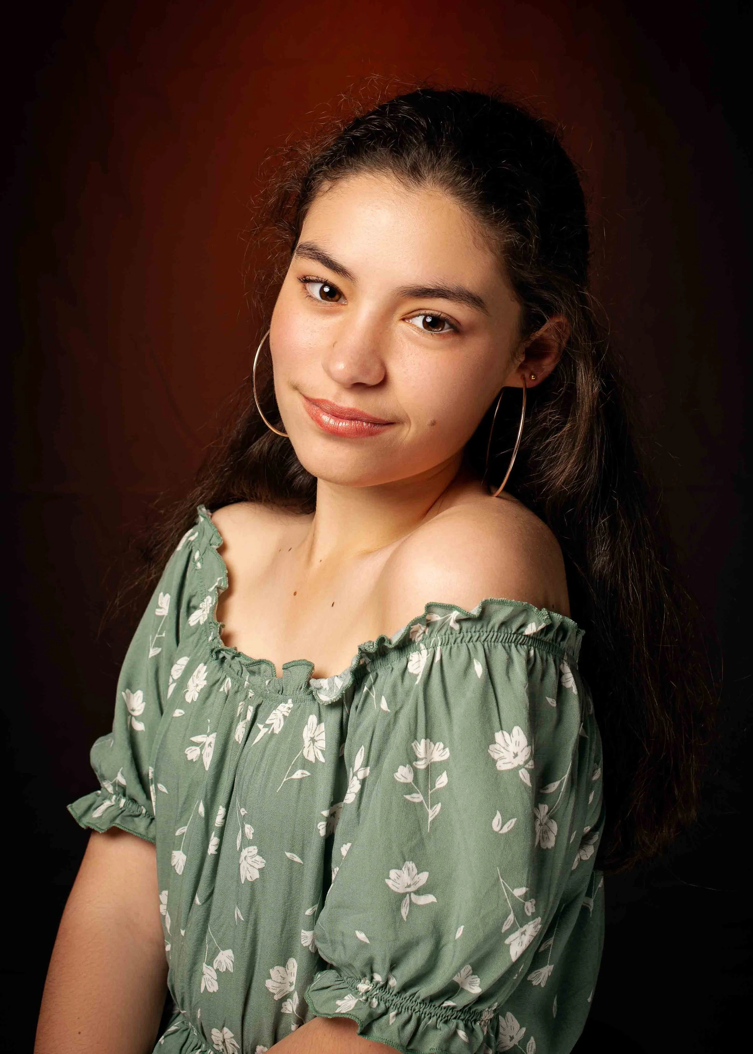 A young woman with long dark hair, hoop earrings, and a soft smile, wearing a green off-the-shoulder top with a white floral pattern, against a dark background.