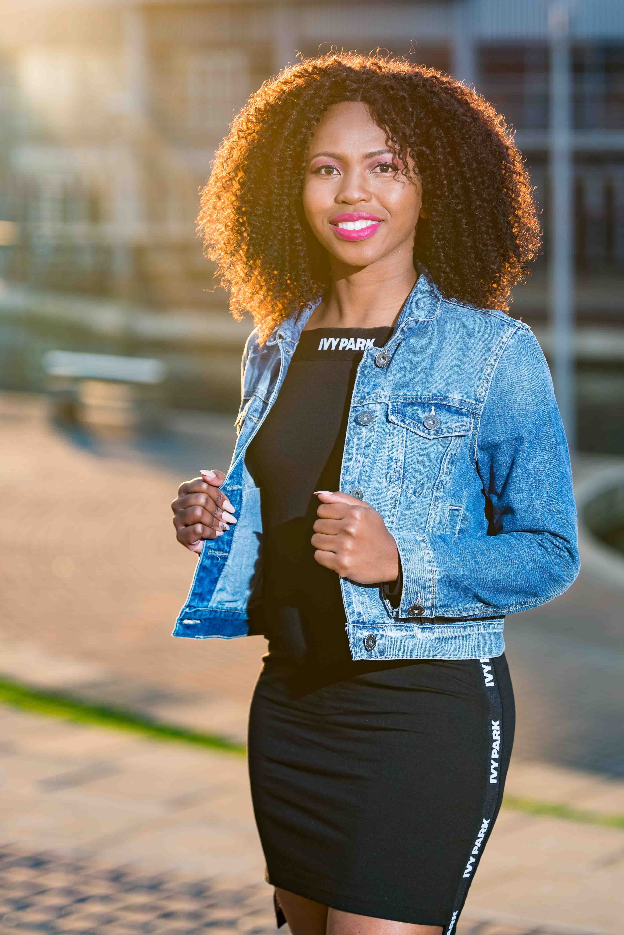 A woman standing outdoors, wearing a denim jacket over a black dress with 'IVY PARK' written on the collar and side, smiling at the camera, with sunlit buildings blurred in the background.