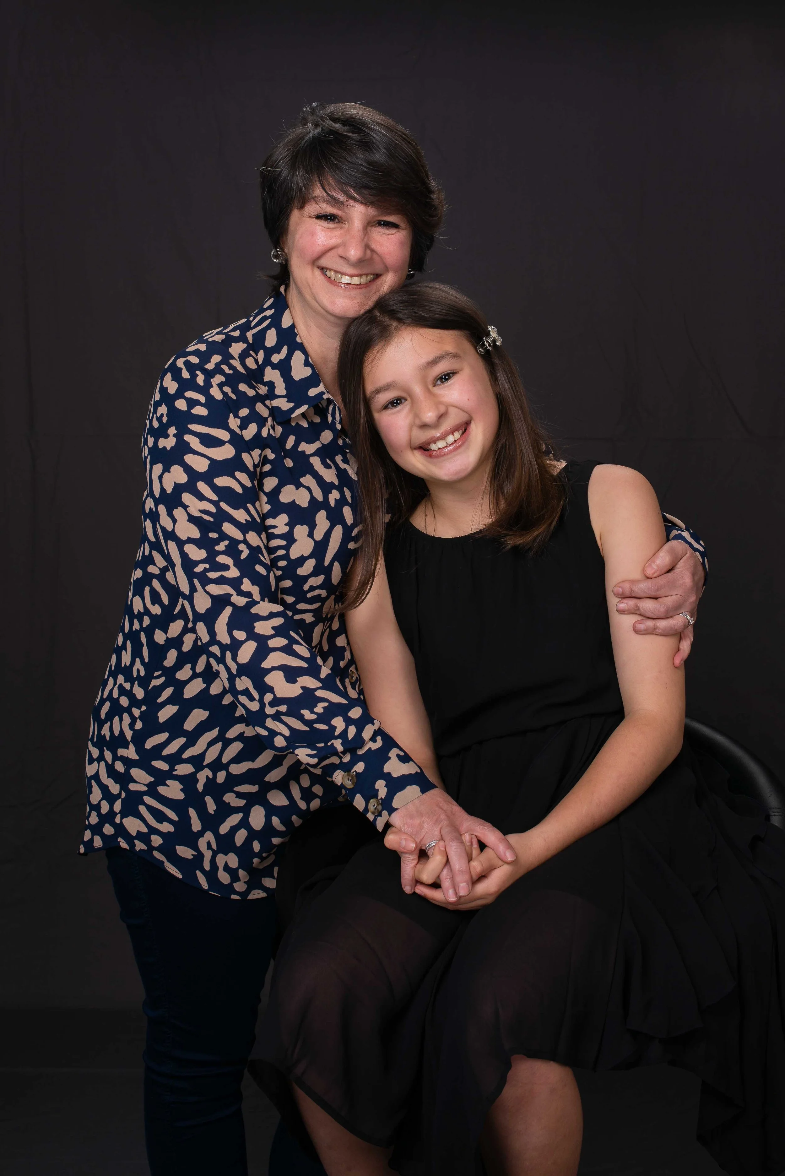 A woman and a young girl smiling and holding hands, with the woman hugging the girl, in front of a plain black backdrop.