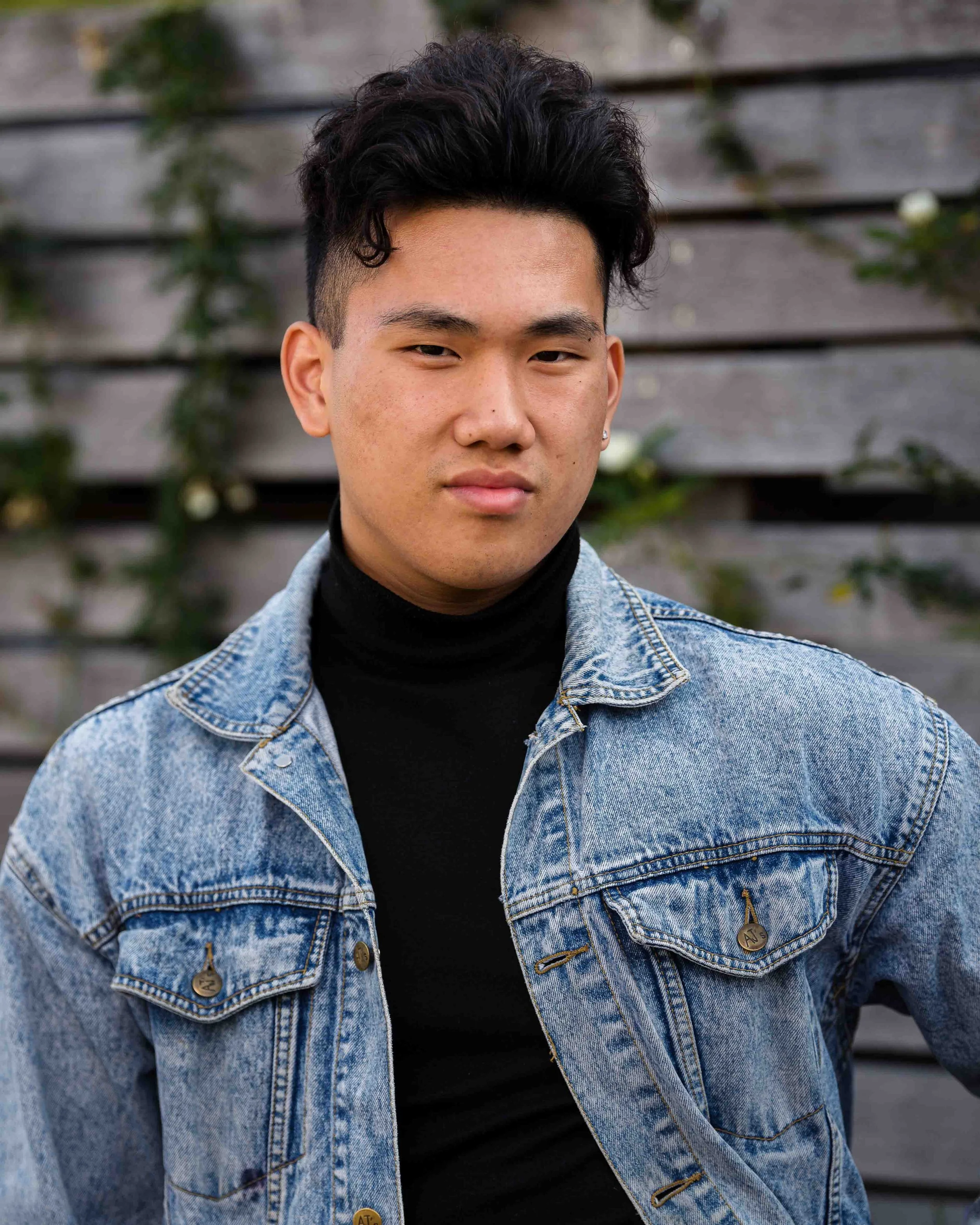 A young man with short black hair wearing a black turtleneck and a denim jacket standing outdoors in front of a wooden fence with plants.