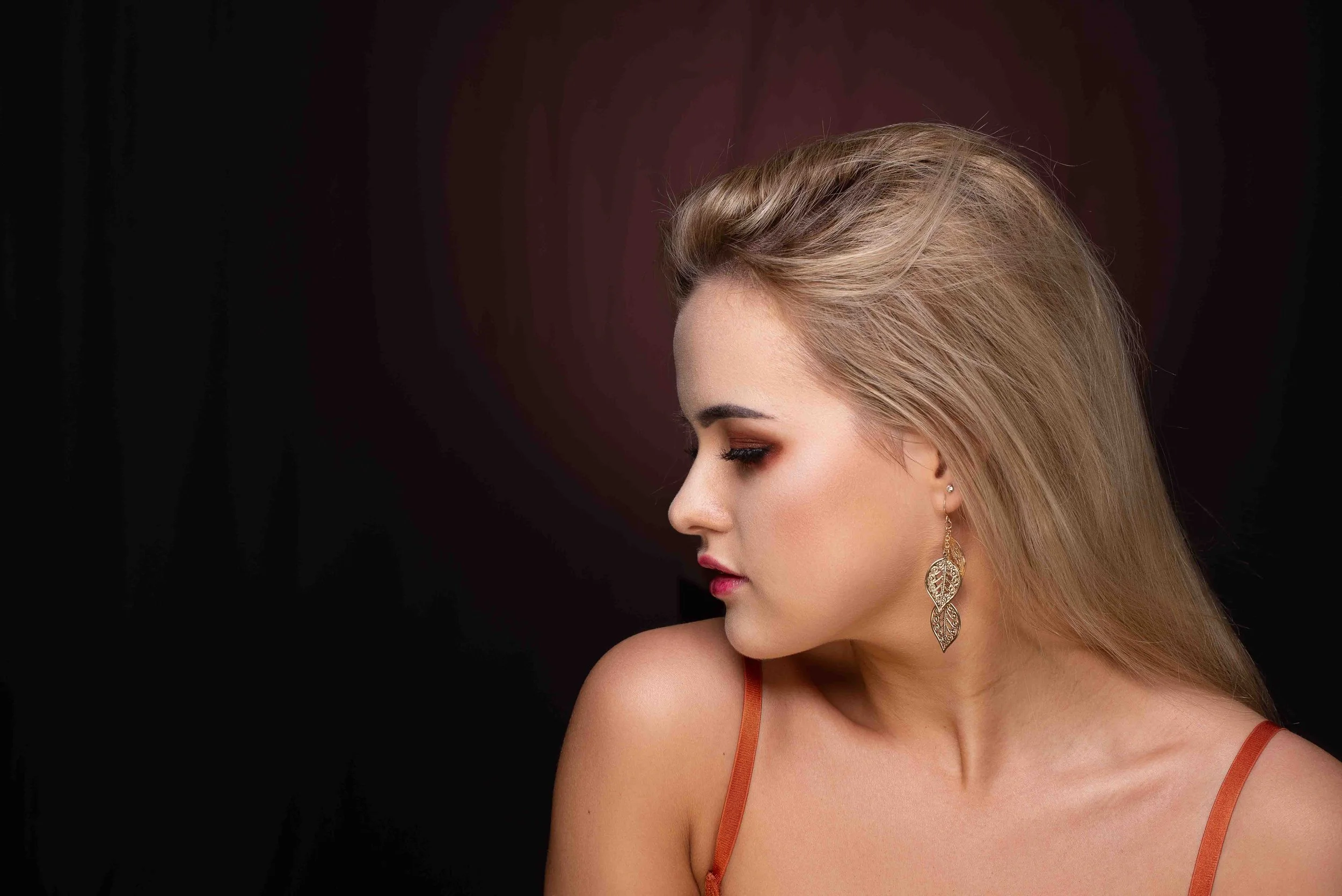 A young woman with blonde hair styled in loose waves, wearing a red strap dress and large leaf-shaped earrings, looking to her left against a dark background.