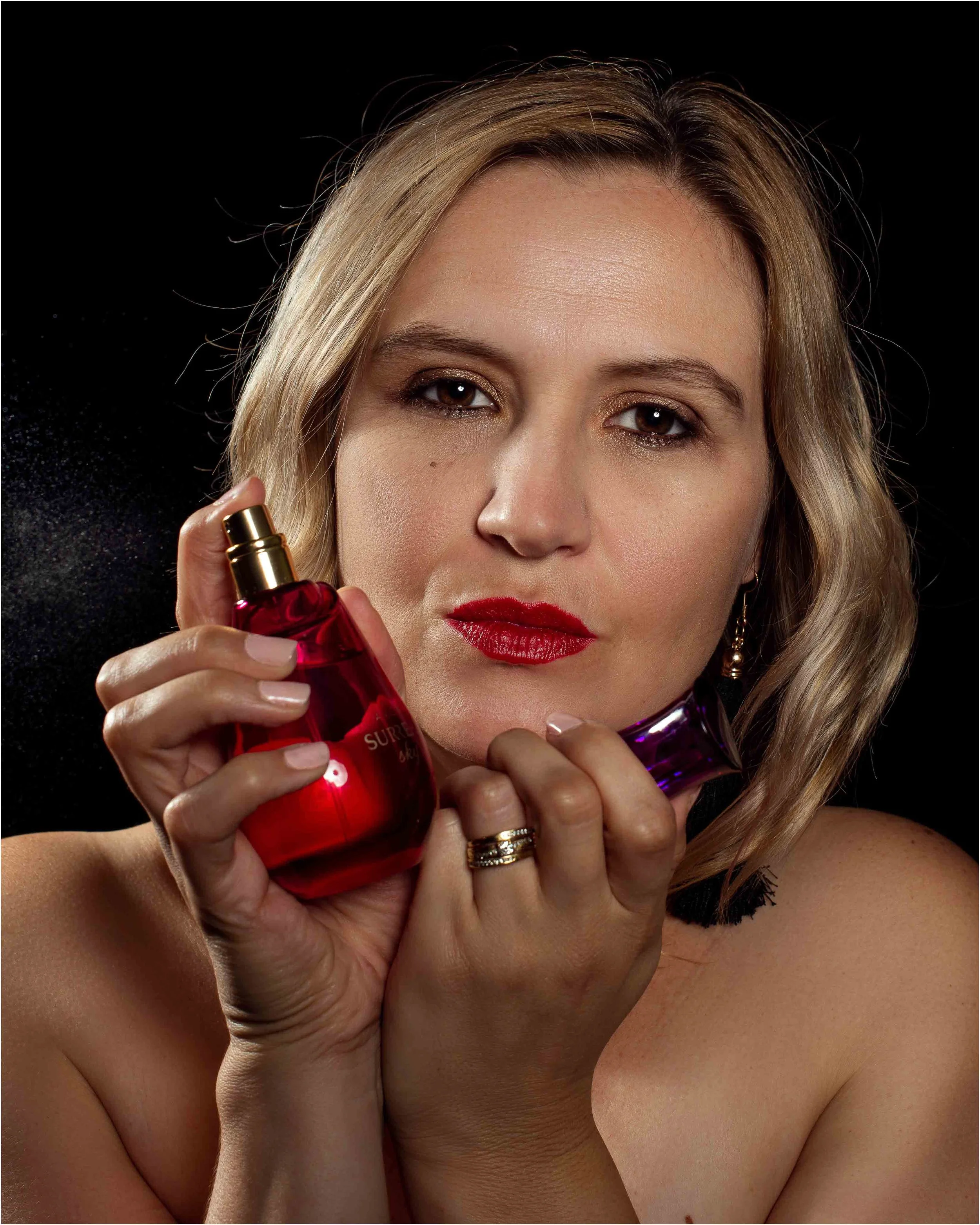 A woman with blonde hair and red lipstick holding two perfume bottles, one red and one purple, against a black background.
