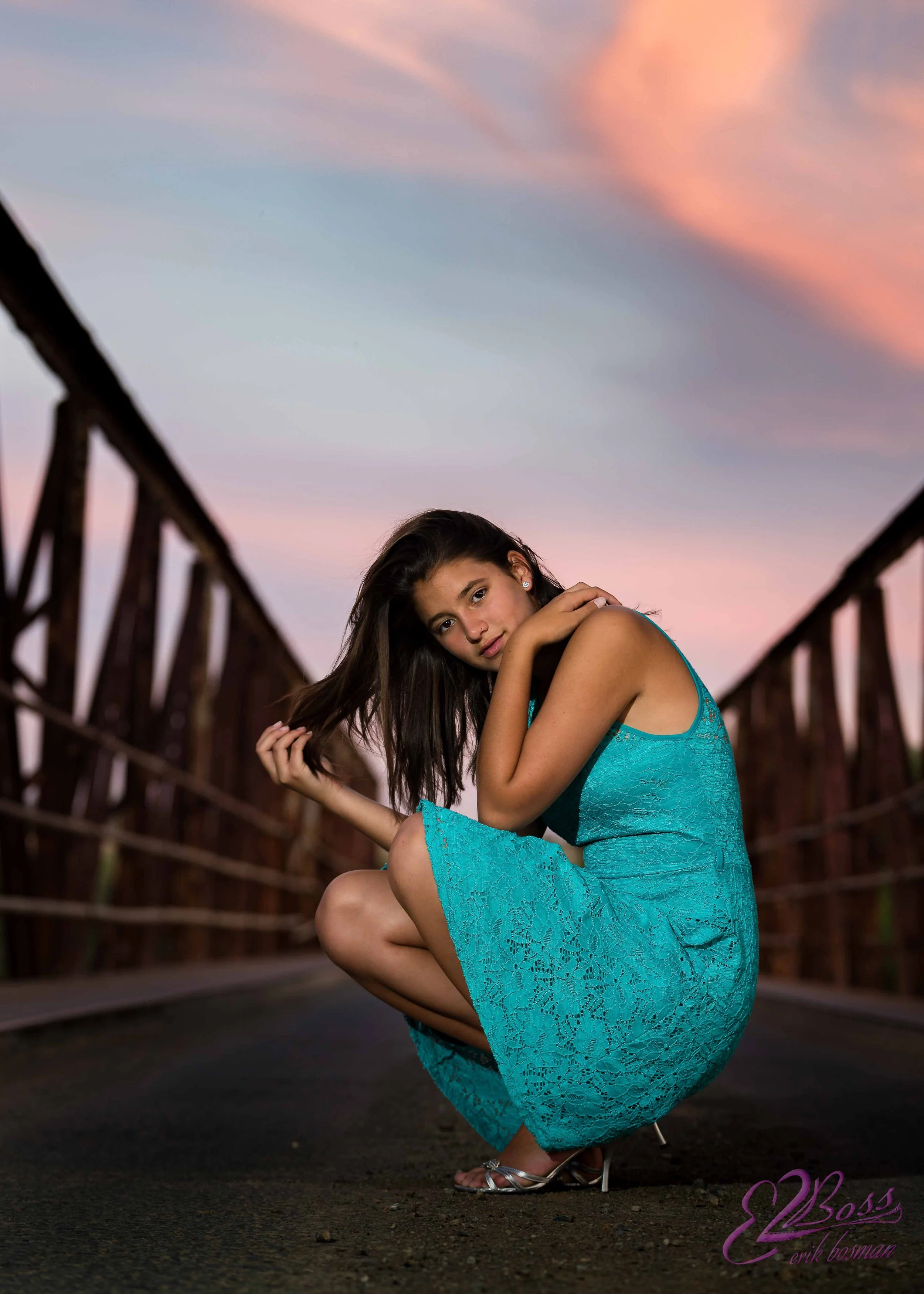 A young woman in a turquoise lace dress squatting on a bridge at sunset, with her hair down and looking at the camera.