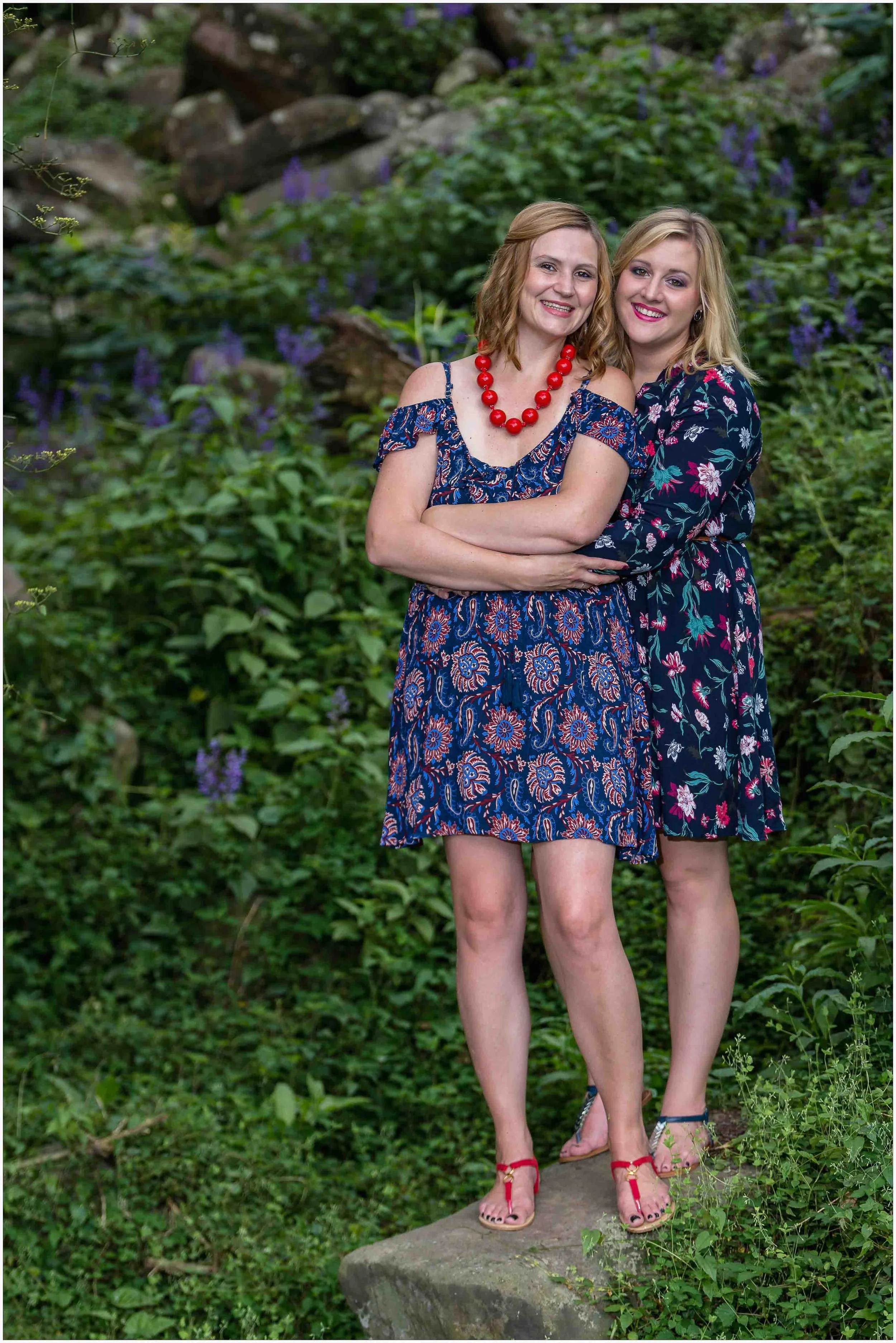 Two women in floral dresses standing outdoors on a rock amid green foliage and purple flowers, smiling and embracing each other.